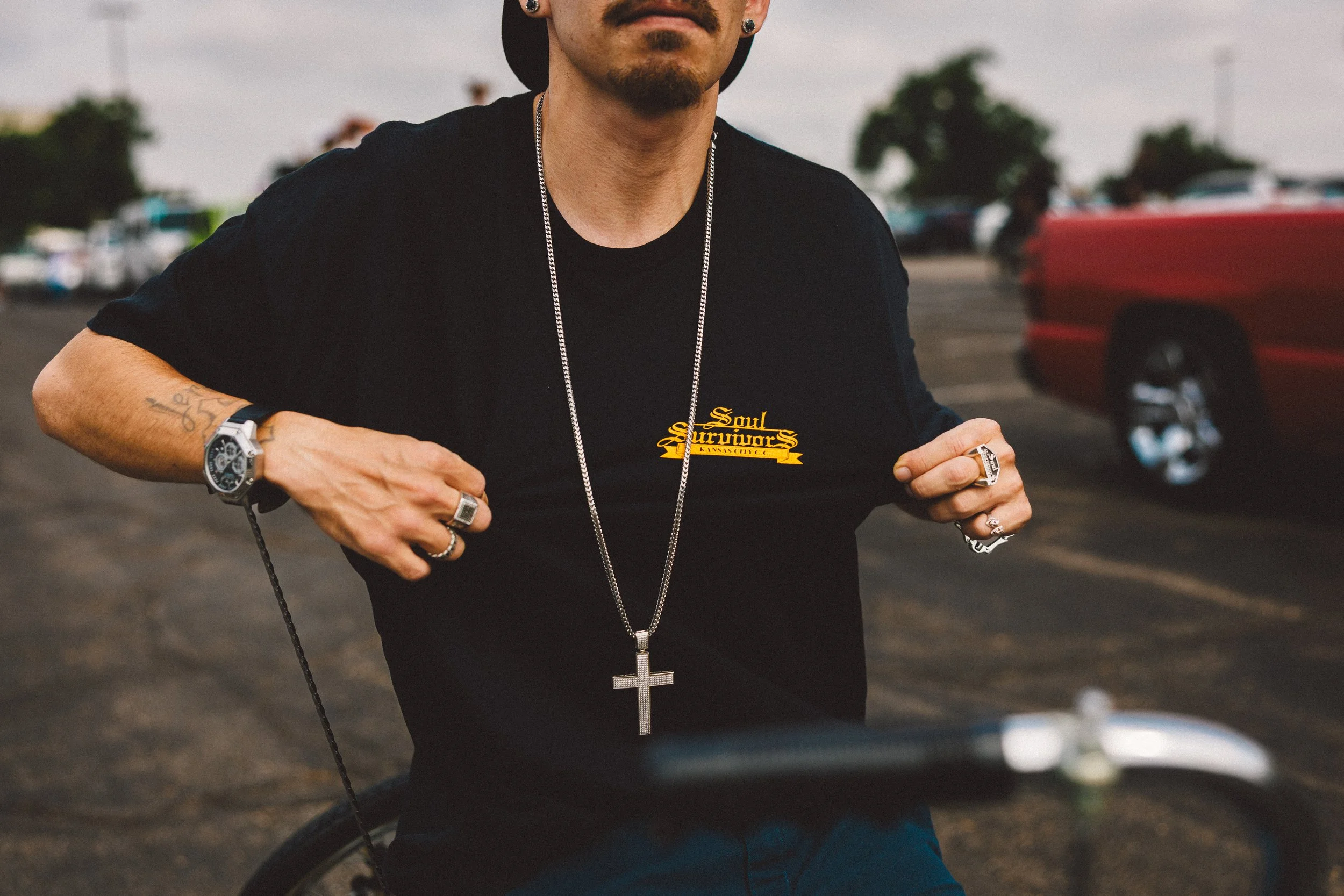 A man wearing a black T-shirt with a yellow logo reading "Soul Survivors" and various jewelry, including a large cross pendant, rings, and rings on his fingers. He is standing outdoors in a parking lot, with cars and trees in the background. 
