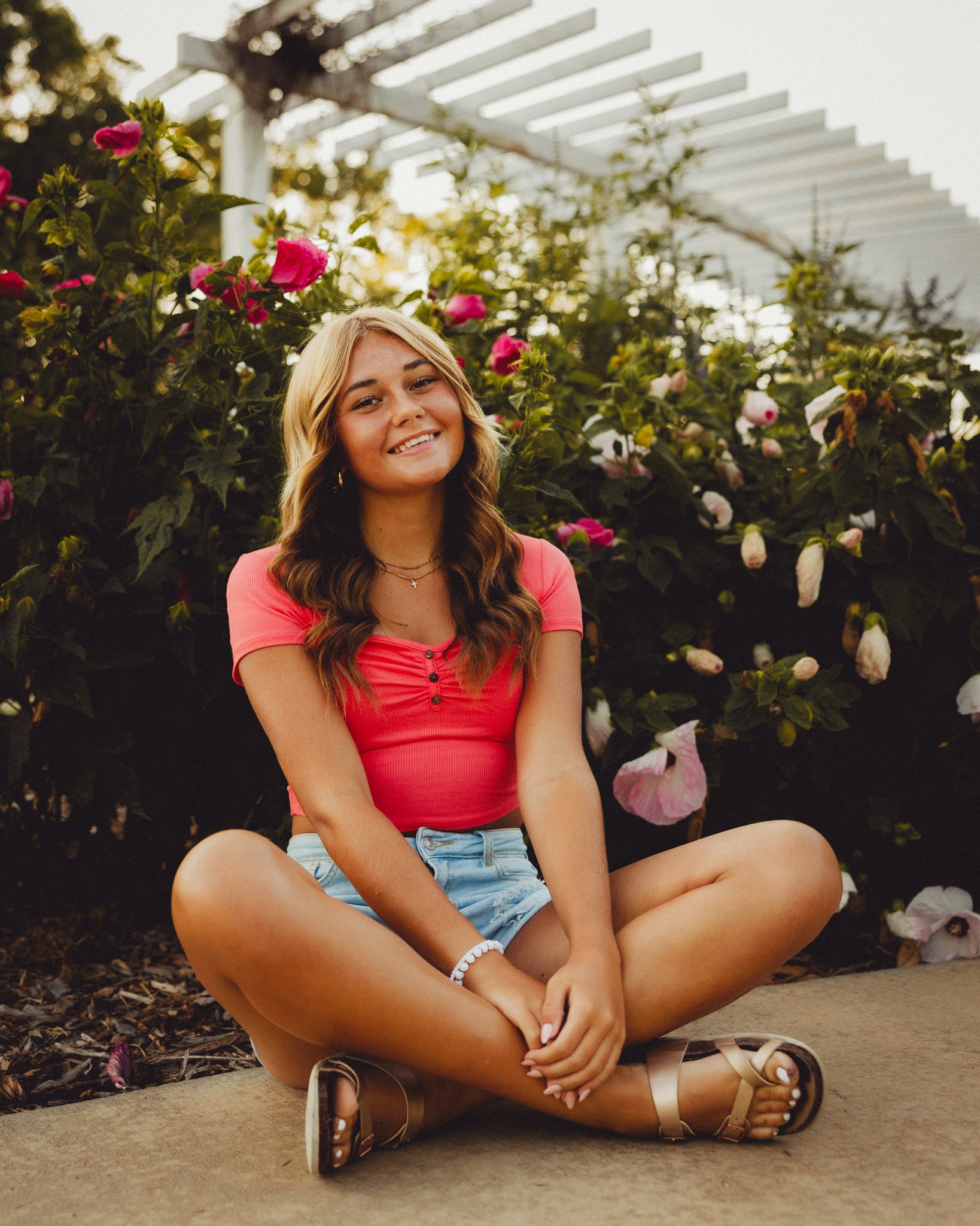 A young woman with long wavy hair, wearing a pink top and denim shorts, sitting cross-legged on a concrete path surrounded by blooming pink and white flowers, with a white wooden pergola in the background during sunset. Shot in Topeka, Kansas. 
