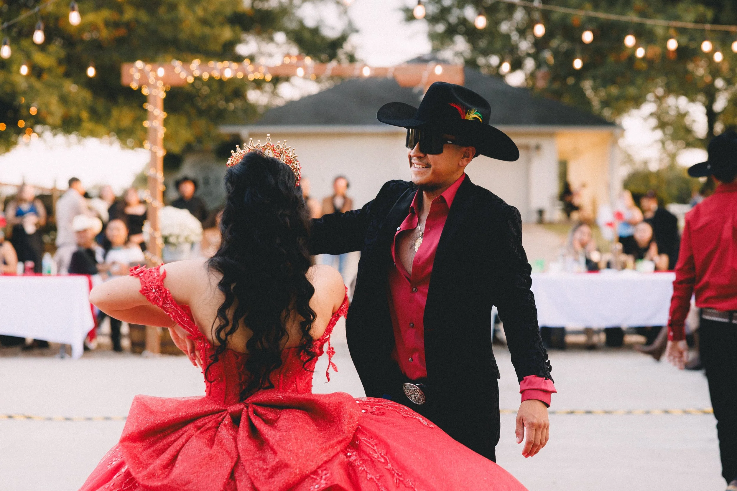 A man and woman are dancing at an outdoor event. The woman is wearing a bright red ball gown with a large bow on the back and a tiara, while the man is dressed in a black cowboy hat, a red shirt, and a black blazer. The background shows other guests,