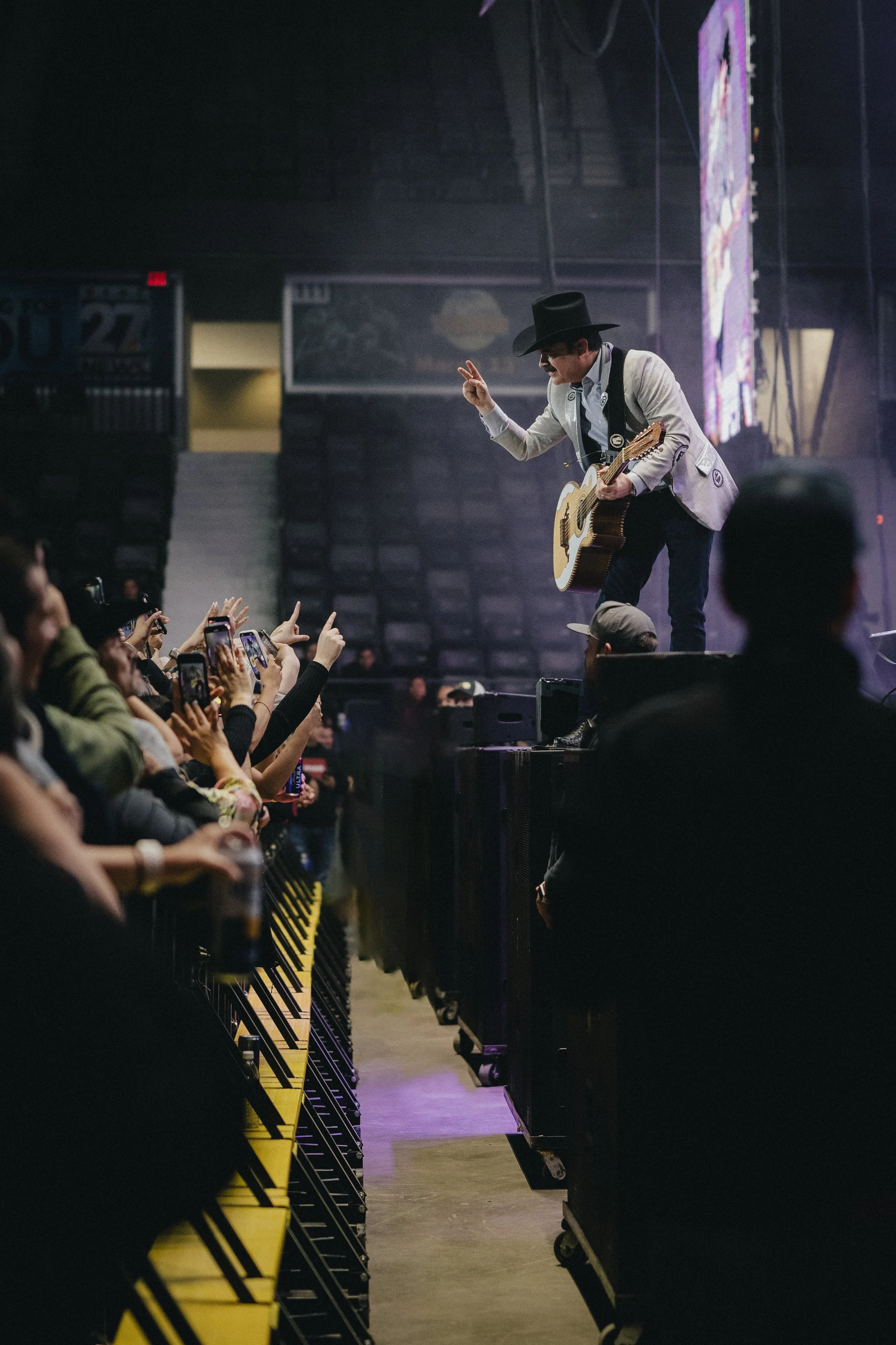 Performer wearing a hat and sunglasses, playing an acoustic guitar on stage at a concert, making a peace sign with his right hand. Audience members are taking pictures and reaching toward the performer. Los Tucanes de Tijuana Stormont Vail Event Cent
