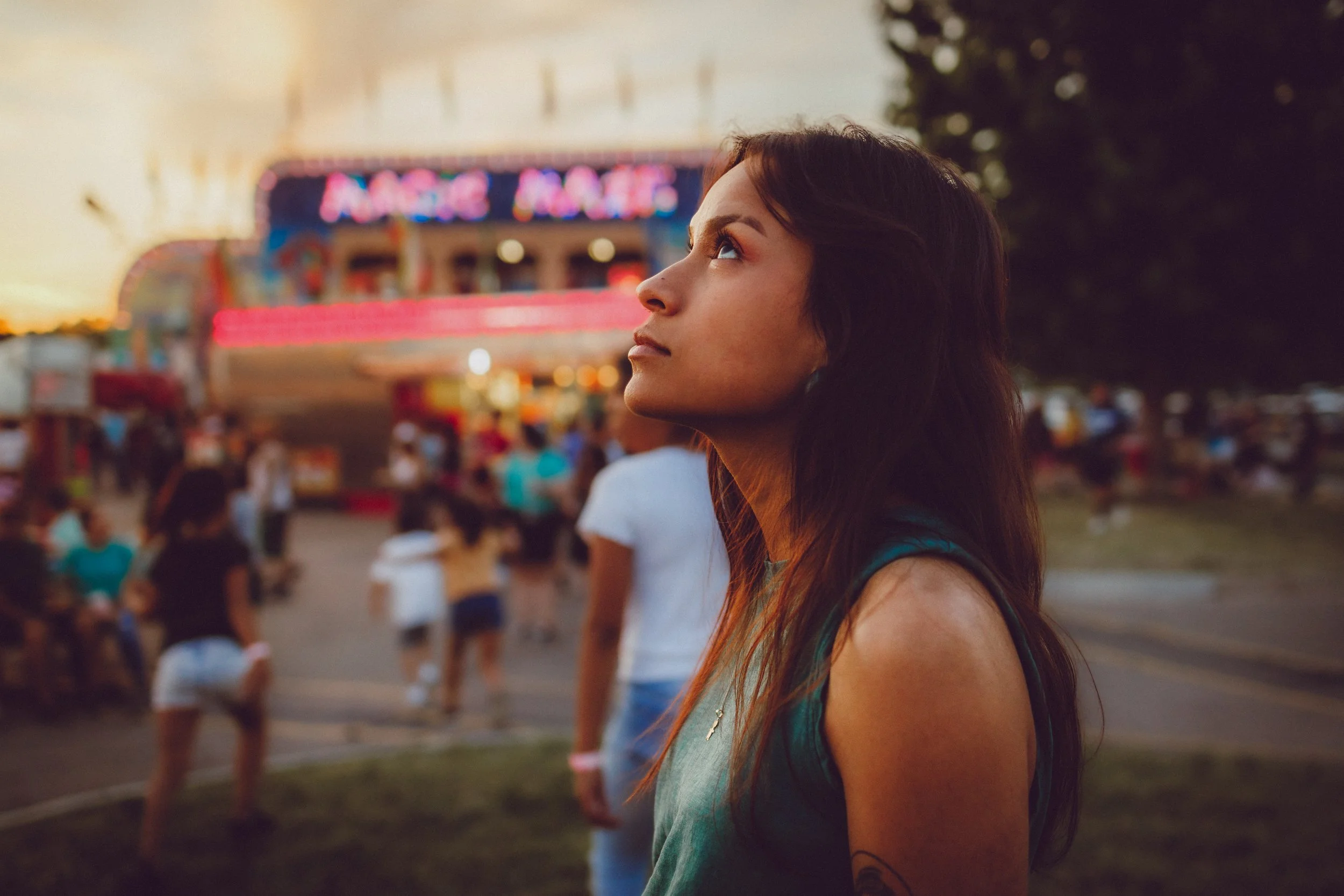 A young woman with long dark hair stands outdoors at a fairground during sunset, looking upward. The fairground has a brightly lit amusement ride and numerous people in the background. 