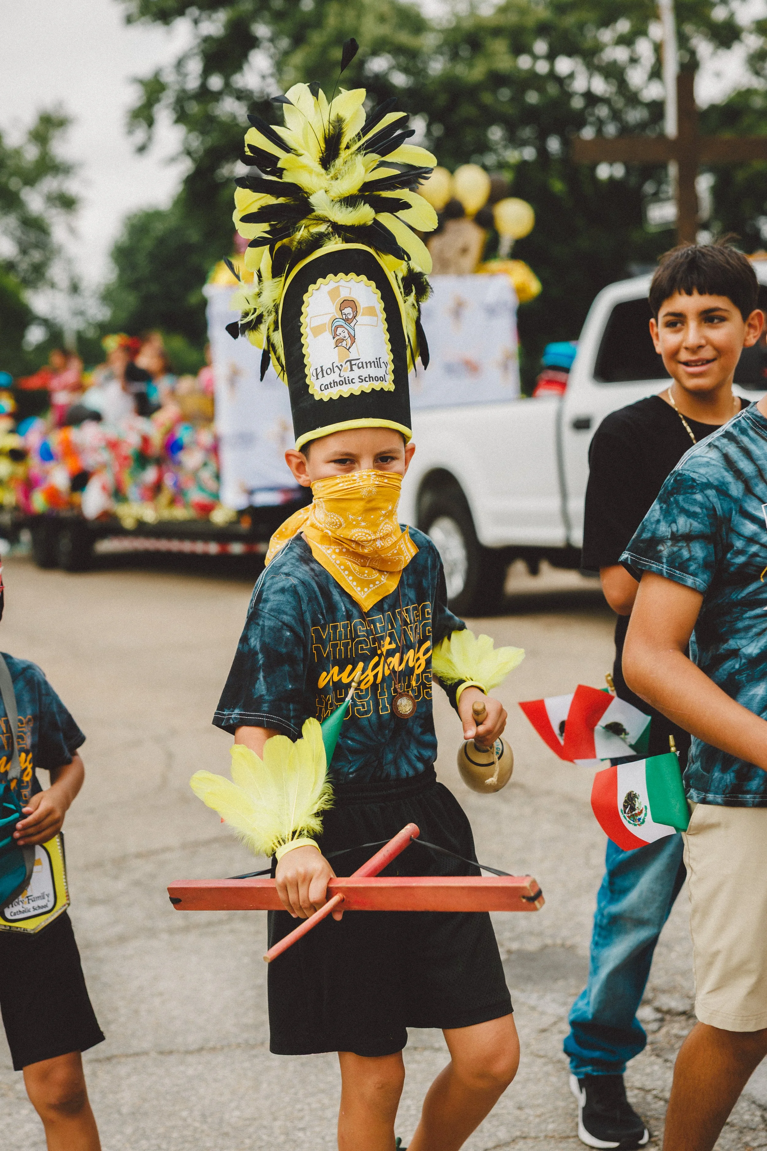 Child dressed in a Detroit Lions shirt, wearing a yellow bandana and a tall headdress with yellow and black feathers, participating in a parade with others holding Mexican flags, a float in the background, and trees and a truck nearby. Shot at Fiesta