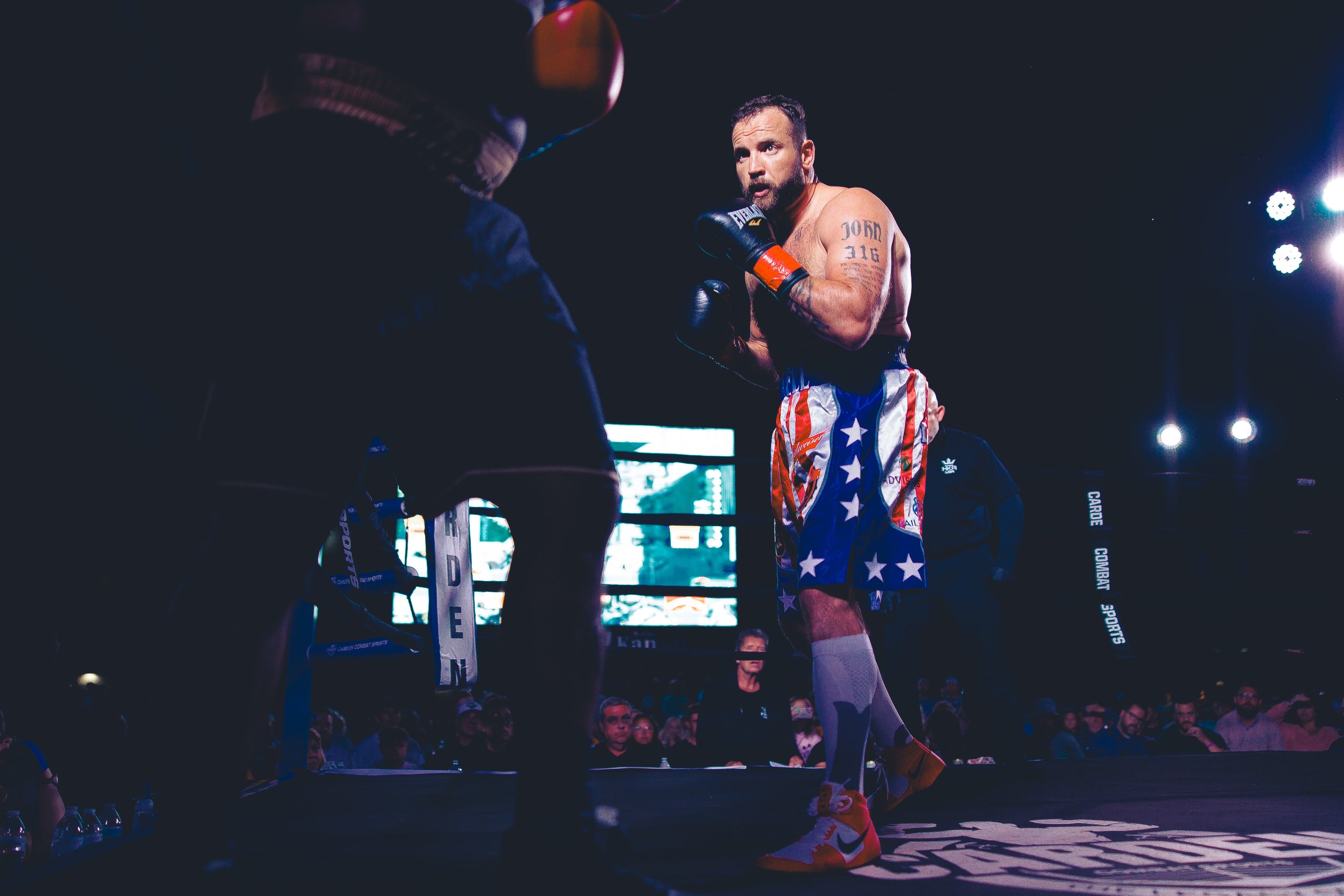A male boxer stands in a boxing ring, facing his opponent, with a serious expression. The boxer is dressed in shorts featuring an American flag pattern, gloves, and sports shoes. The scene is illuminated by bright lights, with a crowd watching in the