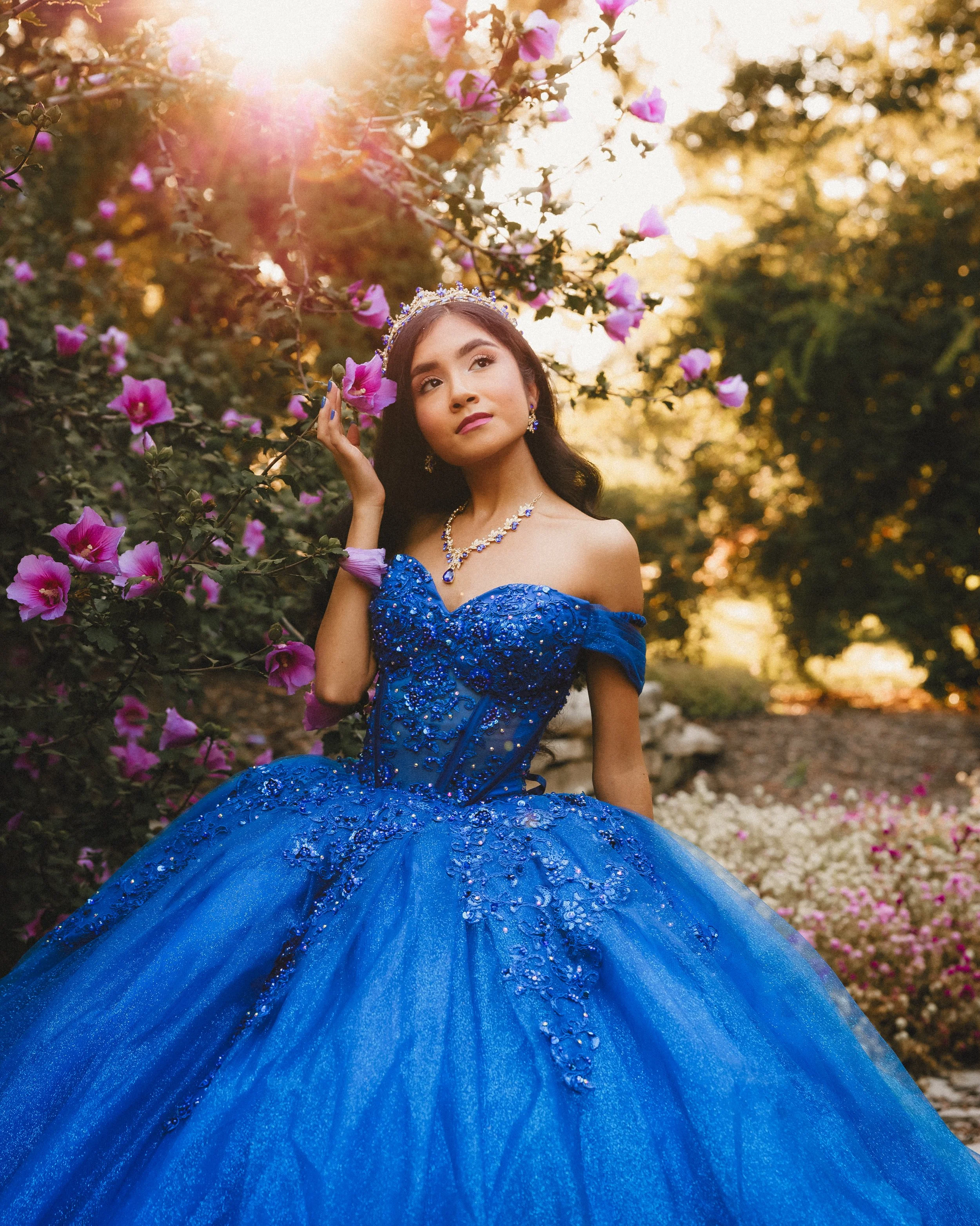 A young woman in an elaborate, off-the-shoulder, royal blue ball gown with intricate beading, standing in a garden among pink flowering bushes, during sunset.