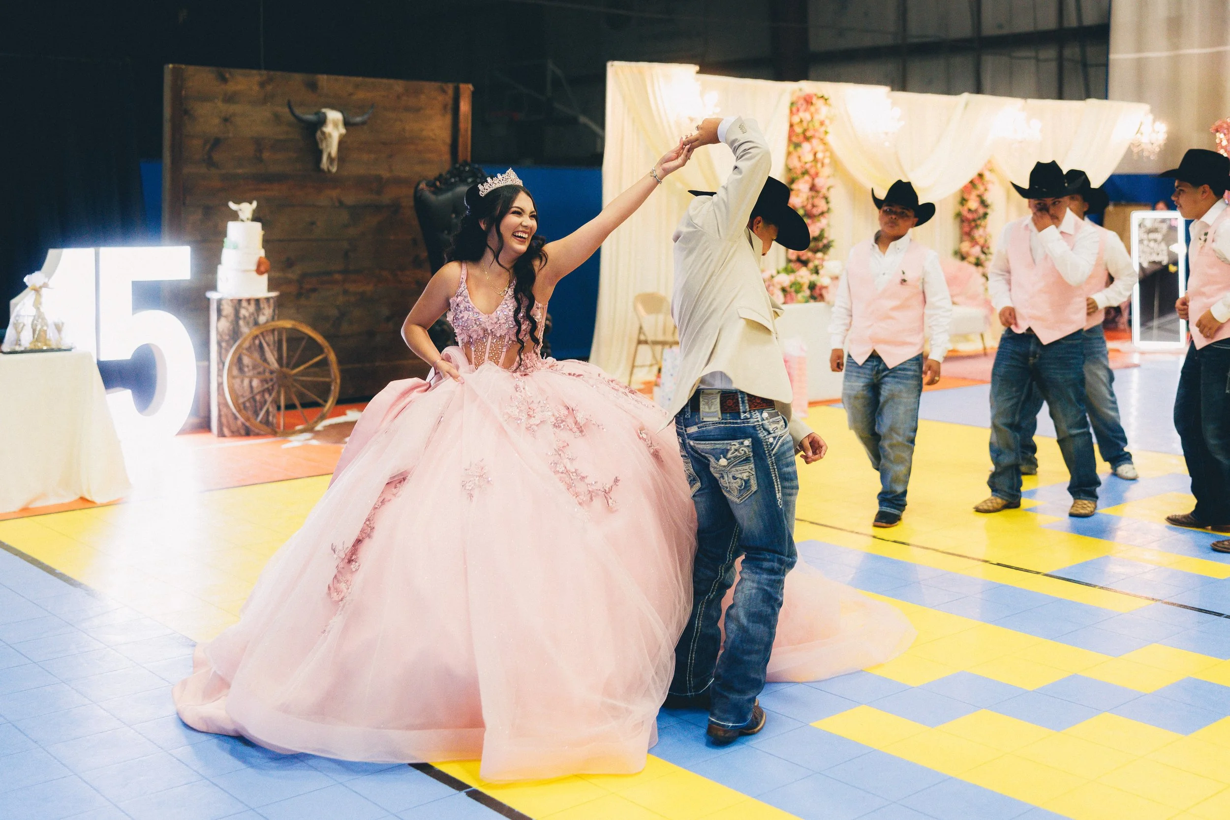 A bride in a pink quinceañera dress dancing with a young man in a white jacket and cowboy hat, surrounded by friends in cowboy attire at a celebration.