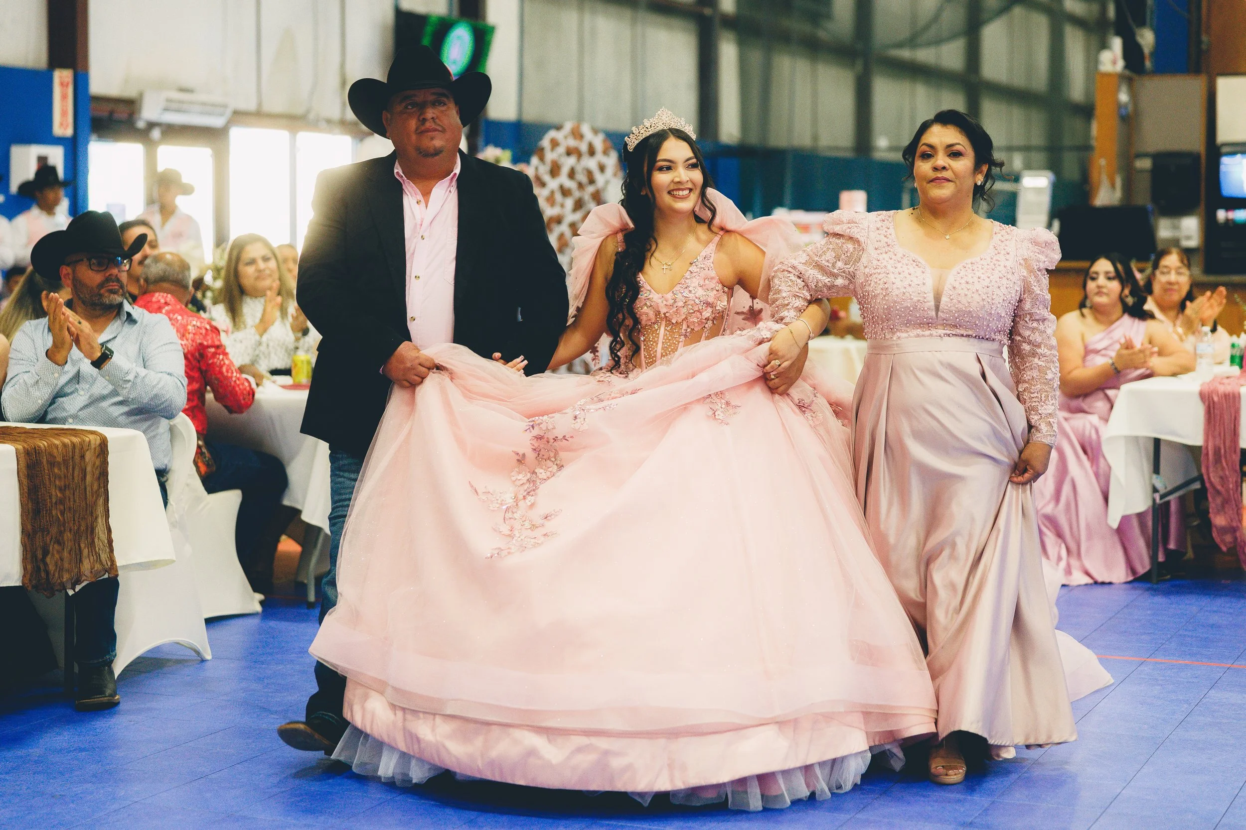 A young woman dressed in a pink quinceañera gown, wearing a tiara, is being escorted by two women and a man at a celebration in a decorated hall. The woman on the right wears a matching pink dress, and the man on the left is in a cowboy hat and black