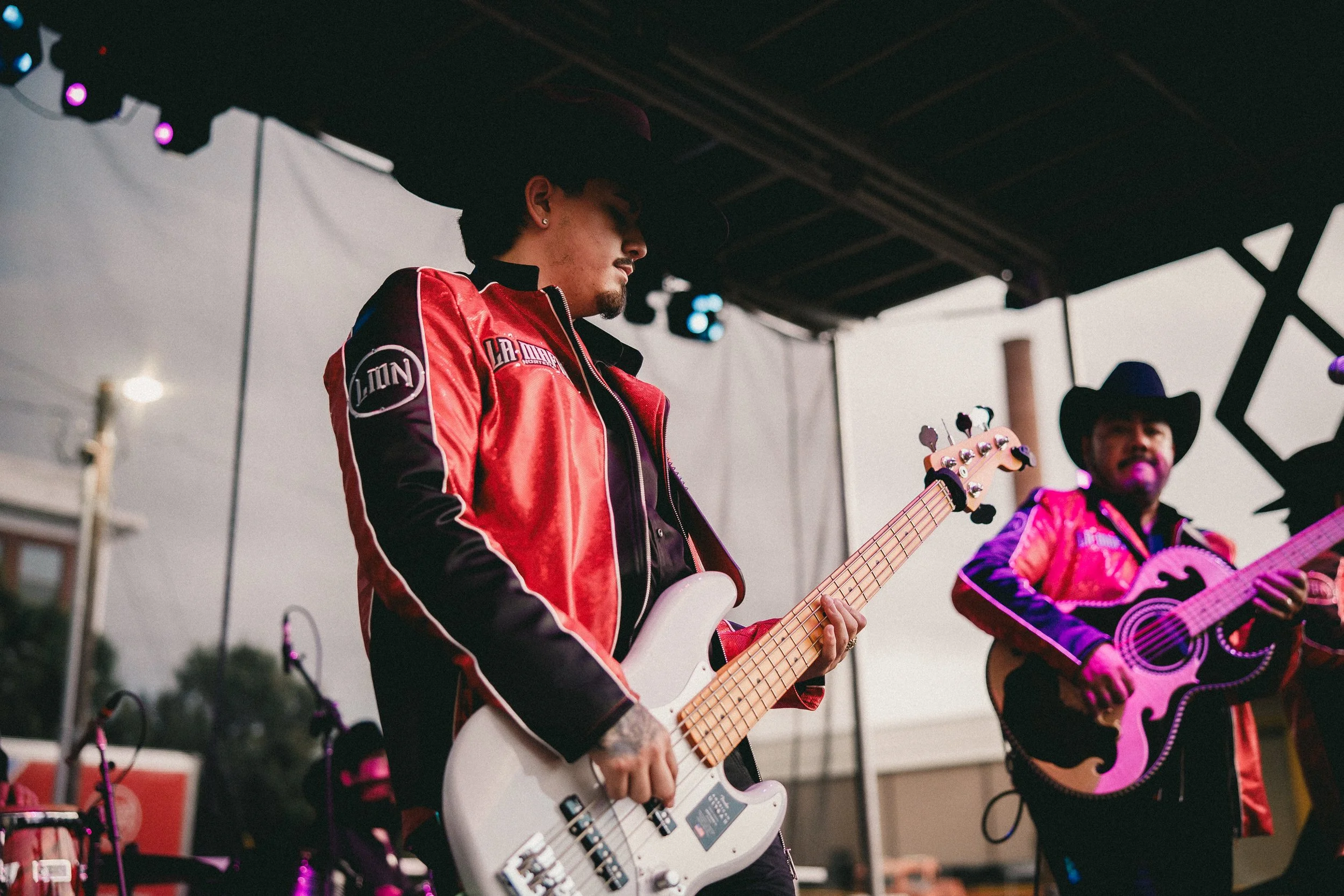 Two musicians performing on an outdoor stage: one playing a bass guitar and the other playing a guitar. Both are wearing Western-style outfits with black hats, red and black jackets. Shot at Fiesta Topeka.