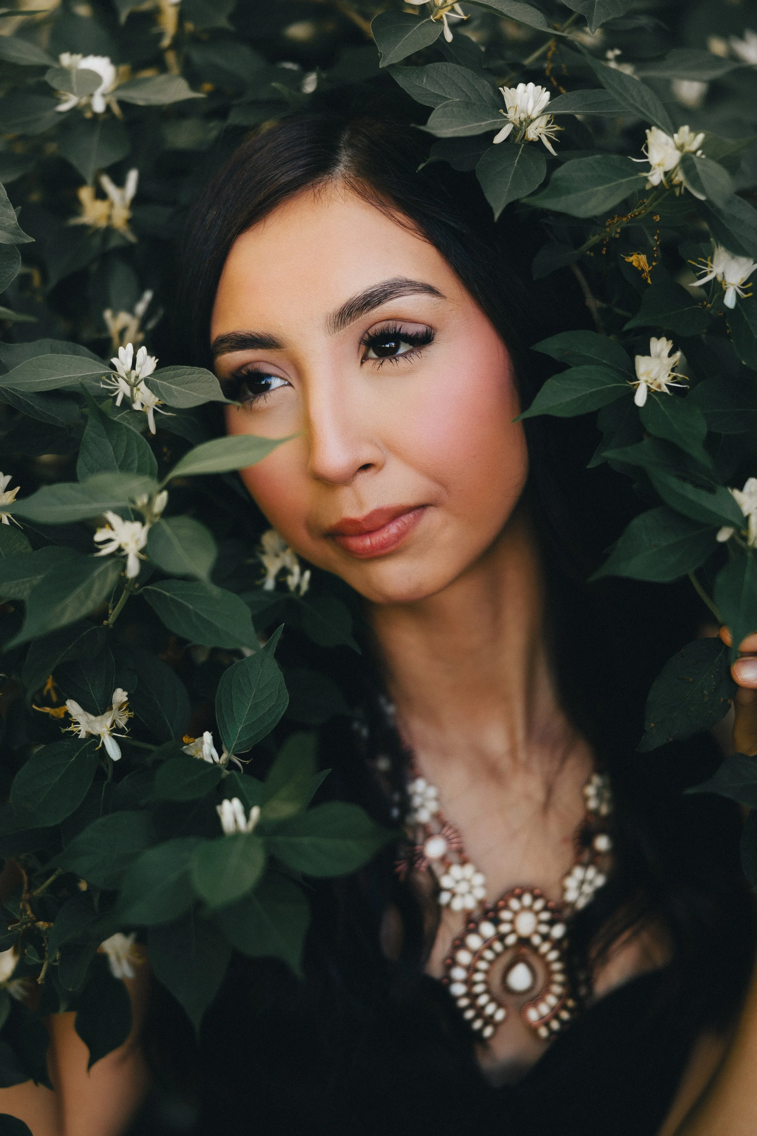 A woman with dark hair and makeup, wearing a statement necklace, is surrounded by green leaves and white flowers.
