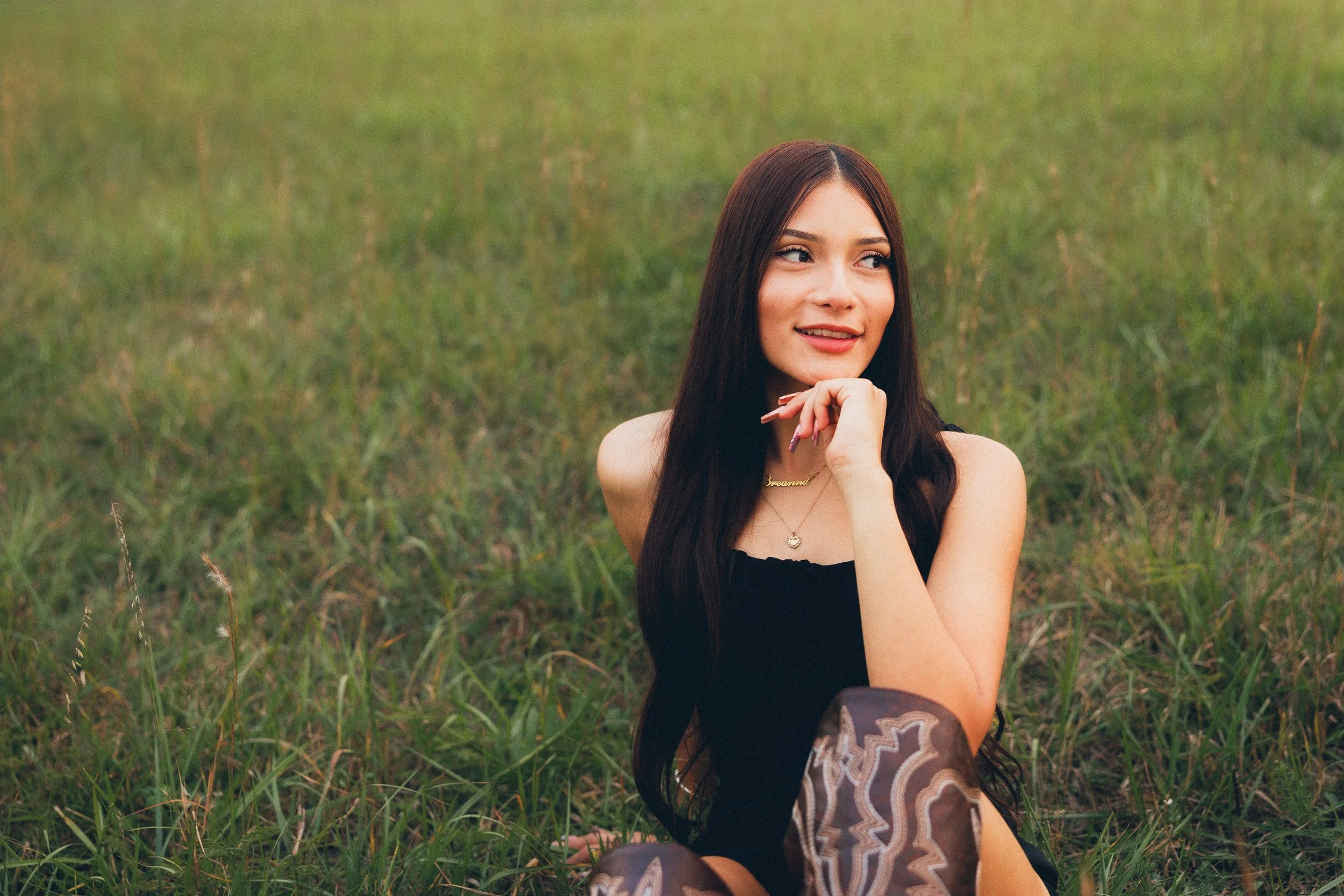 A young woman with long dark hair sitting in a grassy field, wearing a black sleeveless top and patterned tights, smiling and looking to her left.