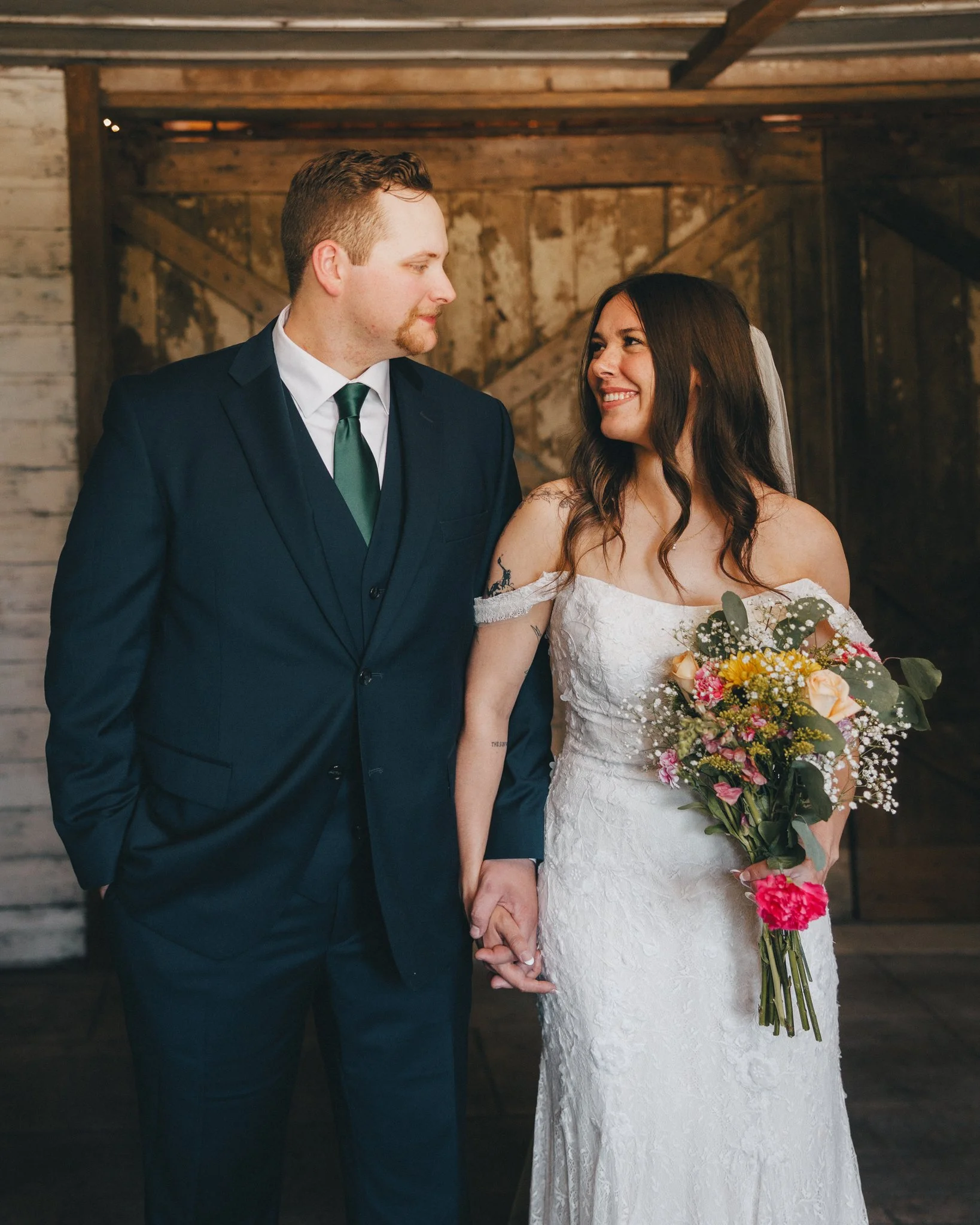 A bride and groom holding hands during their wedding ceremony; the bride is wearing a white lace dress and holding a bouquet, the groom is wearing a dark suit with a white shirt and green tie; they are looking at each other and smiling.