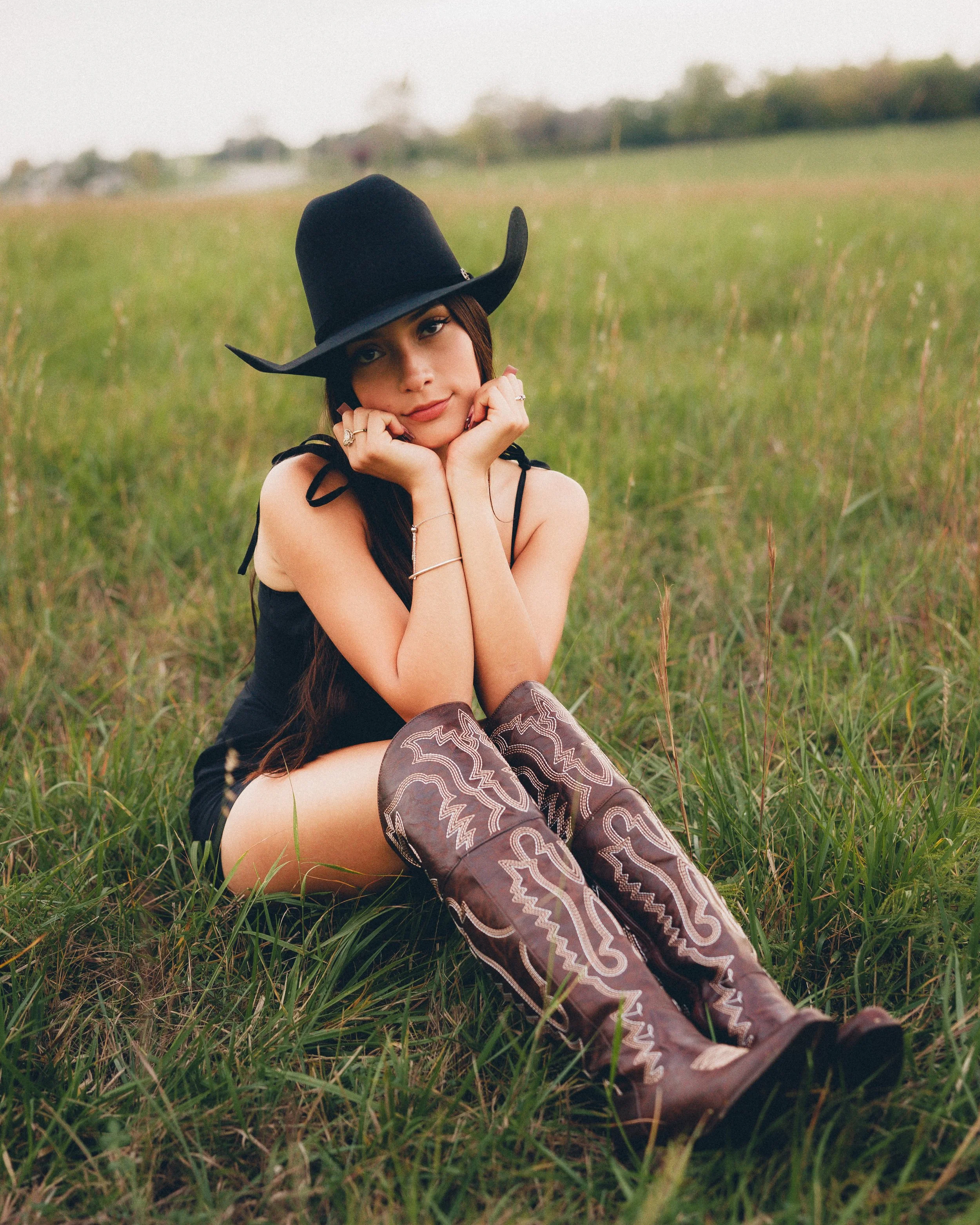 Young woman sitting in a grassy field wearing a black cowboy hat, black dress, and cowboy boots with intricate embroidery, resting her chin on her hands.