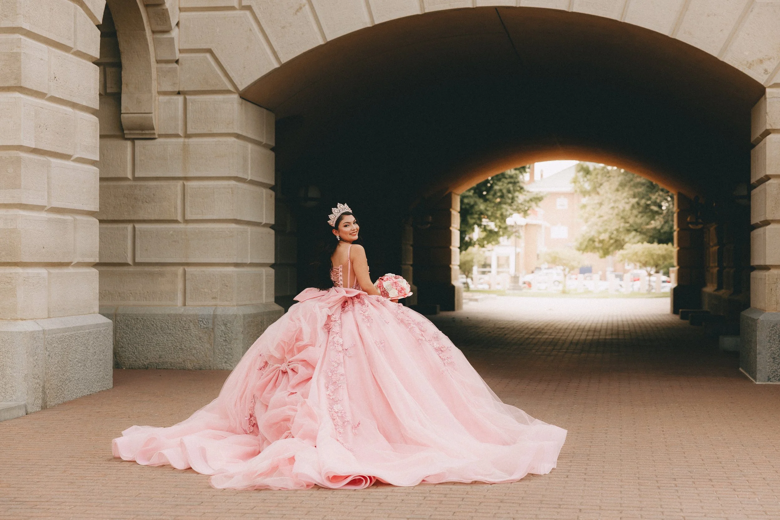 A woman in a pink quinceañera dress with lace details, wearing a tiara, holding a bouquet of pink and white flowers, standing under a stone archway, smiling and looking over her shoulder. Shot in topeka, Kansas. 