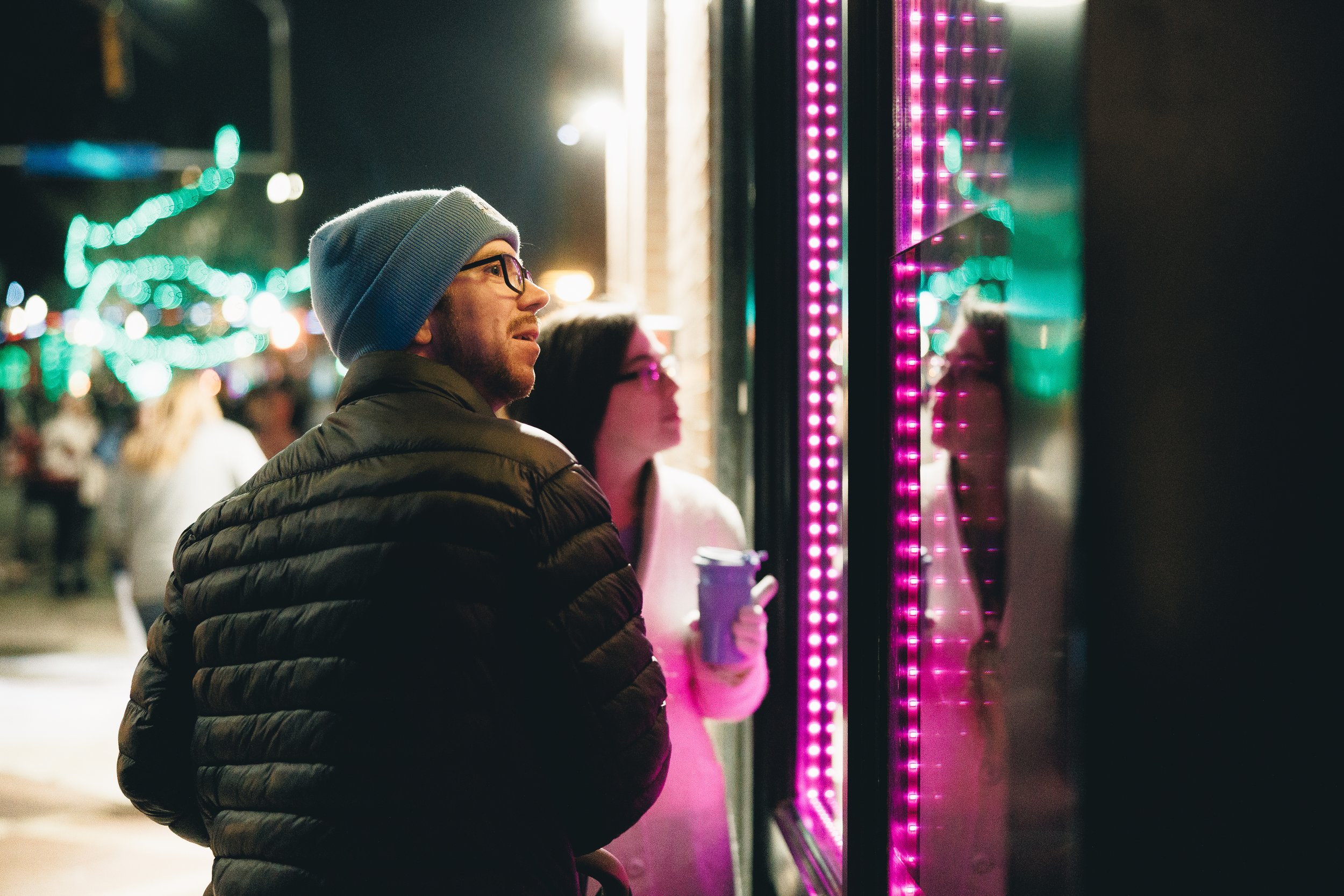 A man and woman standing outside at night, looking at a brightly lit neon sign or display, with other people and colorful lights in the background. Downtown Topeka, Kansas Miracle on Kansas Ave Parade 2025.