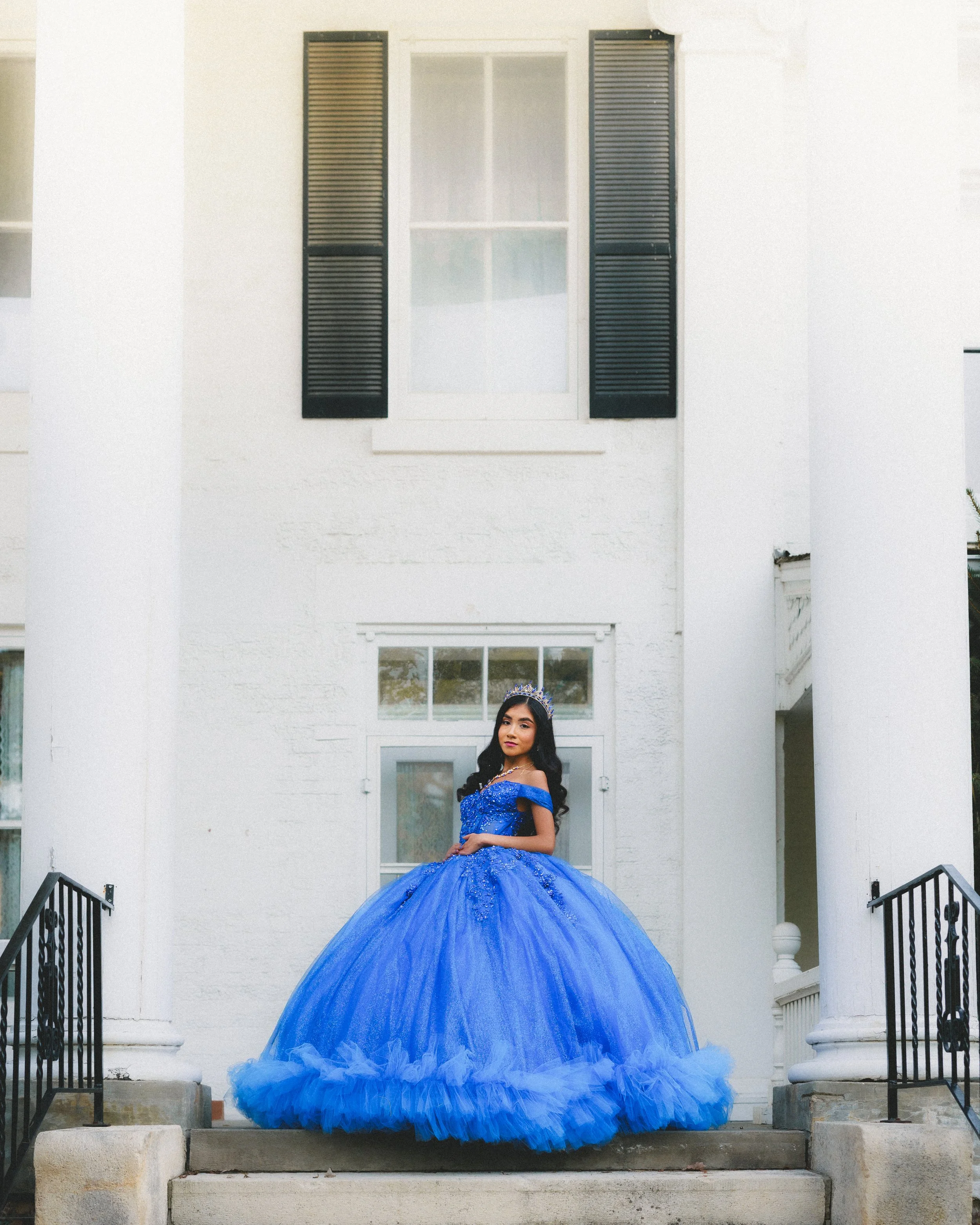 A young woman in a blue ball gown with a tiara standing on the steps of a white building.