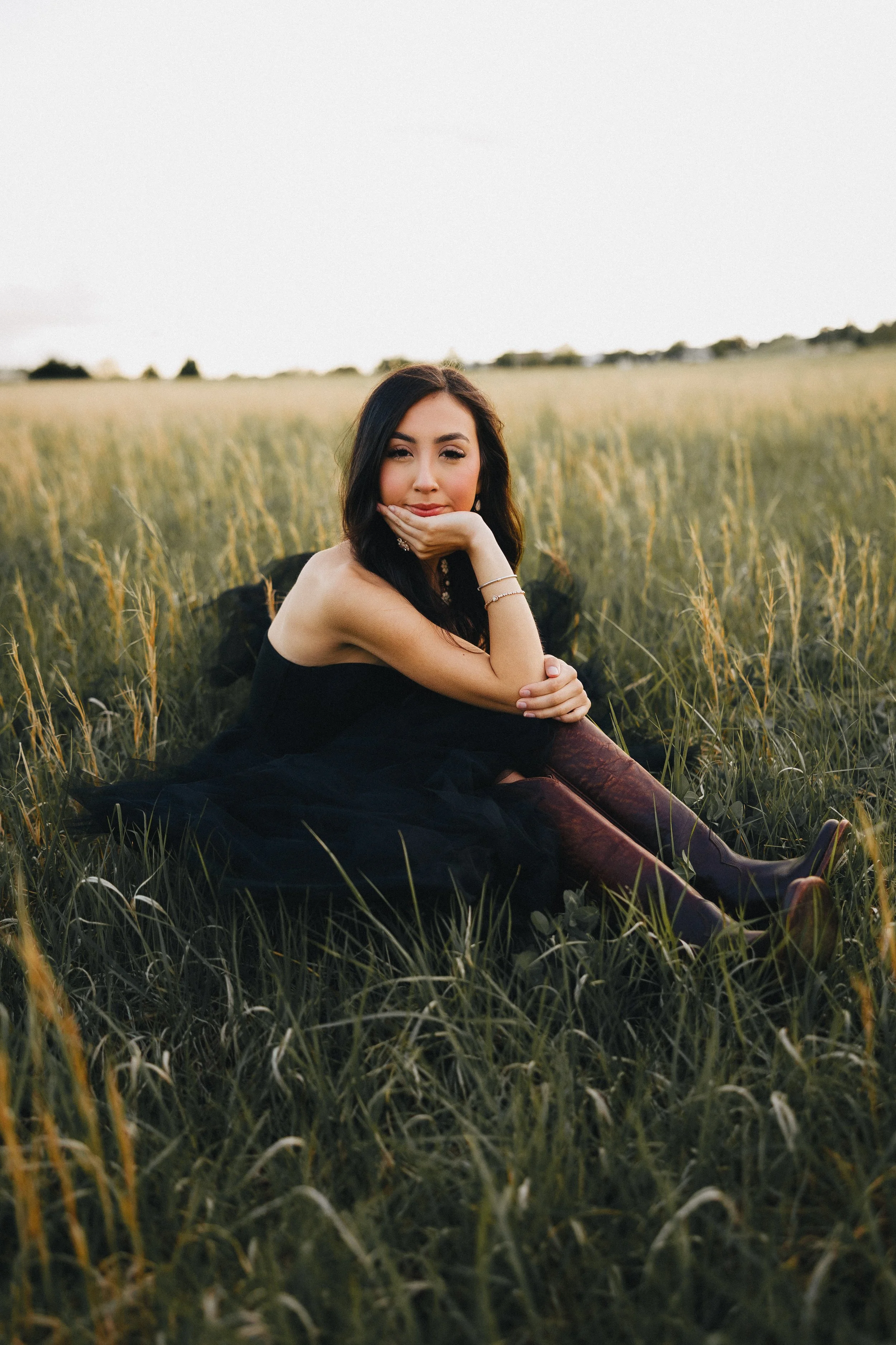 A woman in a black dress and maroon knee-high boots sitting in a grassy field, resting her chin on her hand, with long dark hair, and a serene expression.