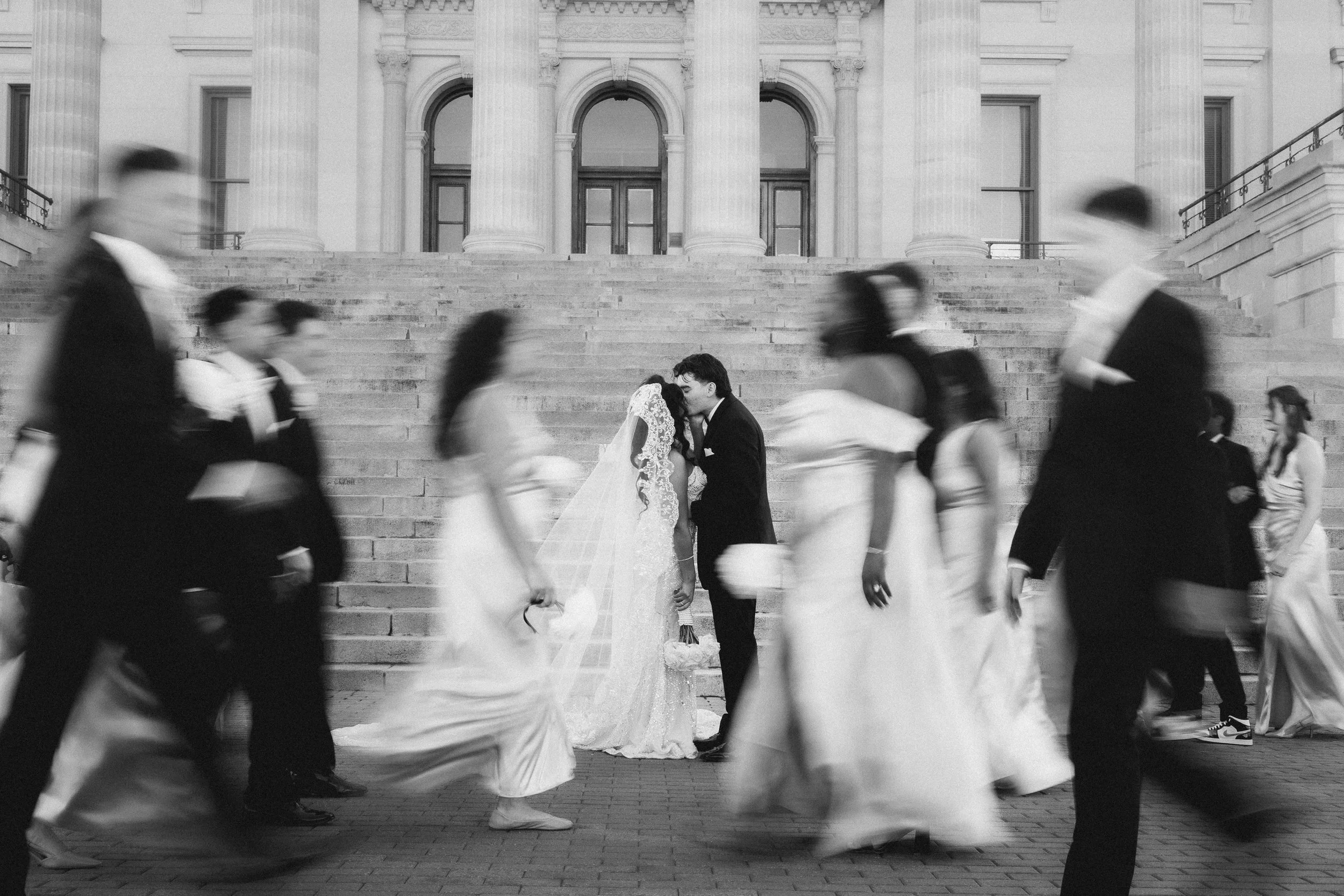 A black and white photo of a bride and groom kissing on the steps of a grand historic building, surrounded by people walking past.