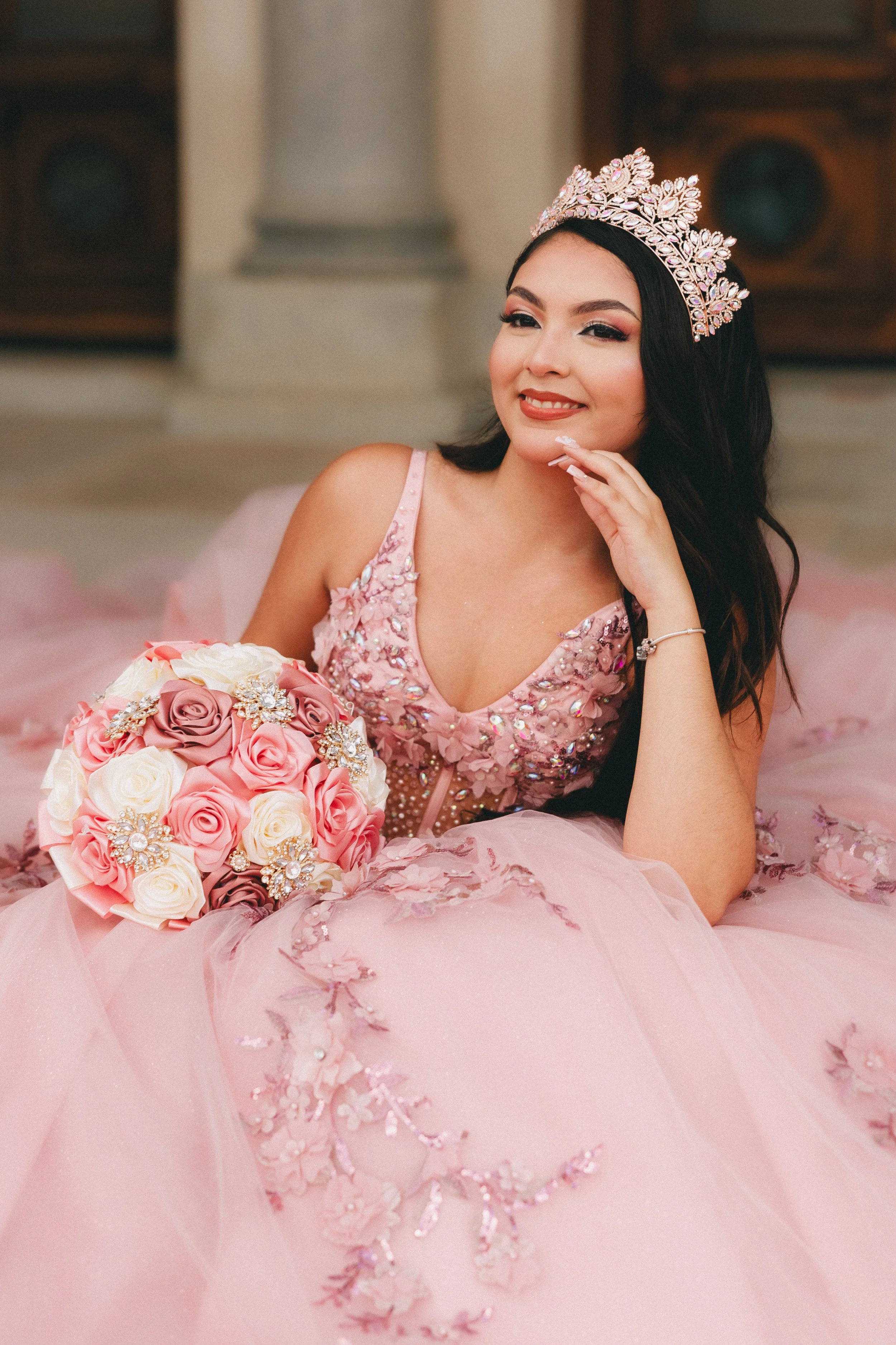 A woman dressed in a pink quinceañera dress with floral embellishments, wearing a tiara, and holding a bouquet of pink and white roses, lying indoors on a pink tulle fabric. Shot in Topeka, Kansas. 