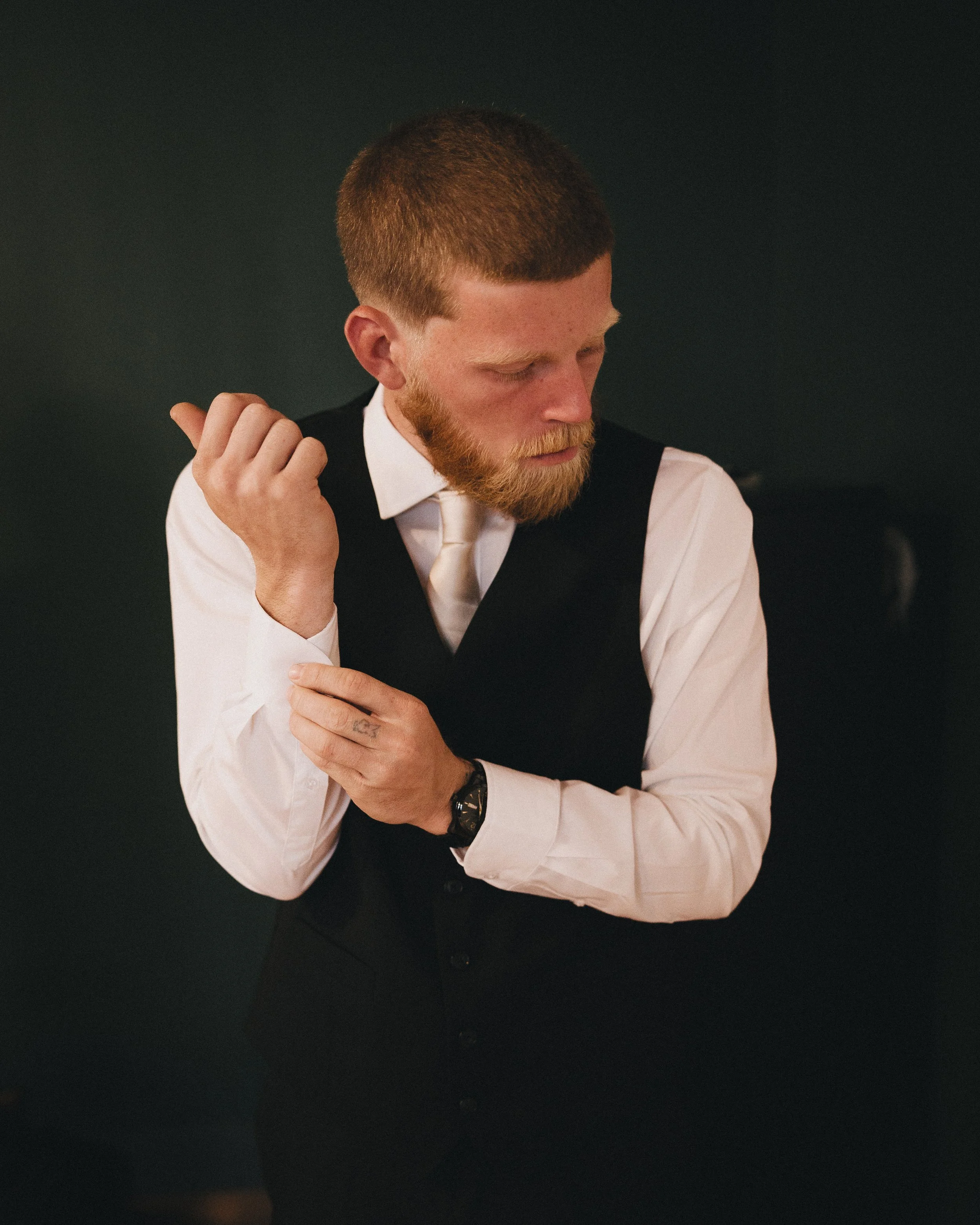 A man with red hair and beard adjusting his cufflink, wearing a white shirt, black vest, and tie, against a dark background. Shot at Topeka Kansas venue The Beacon.