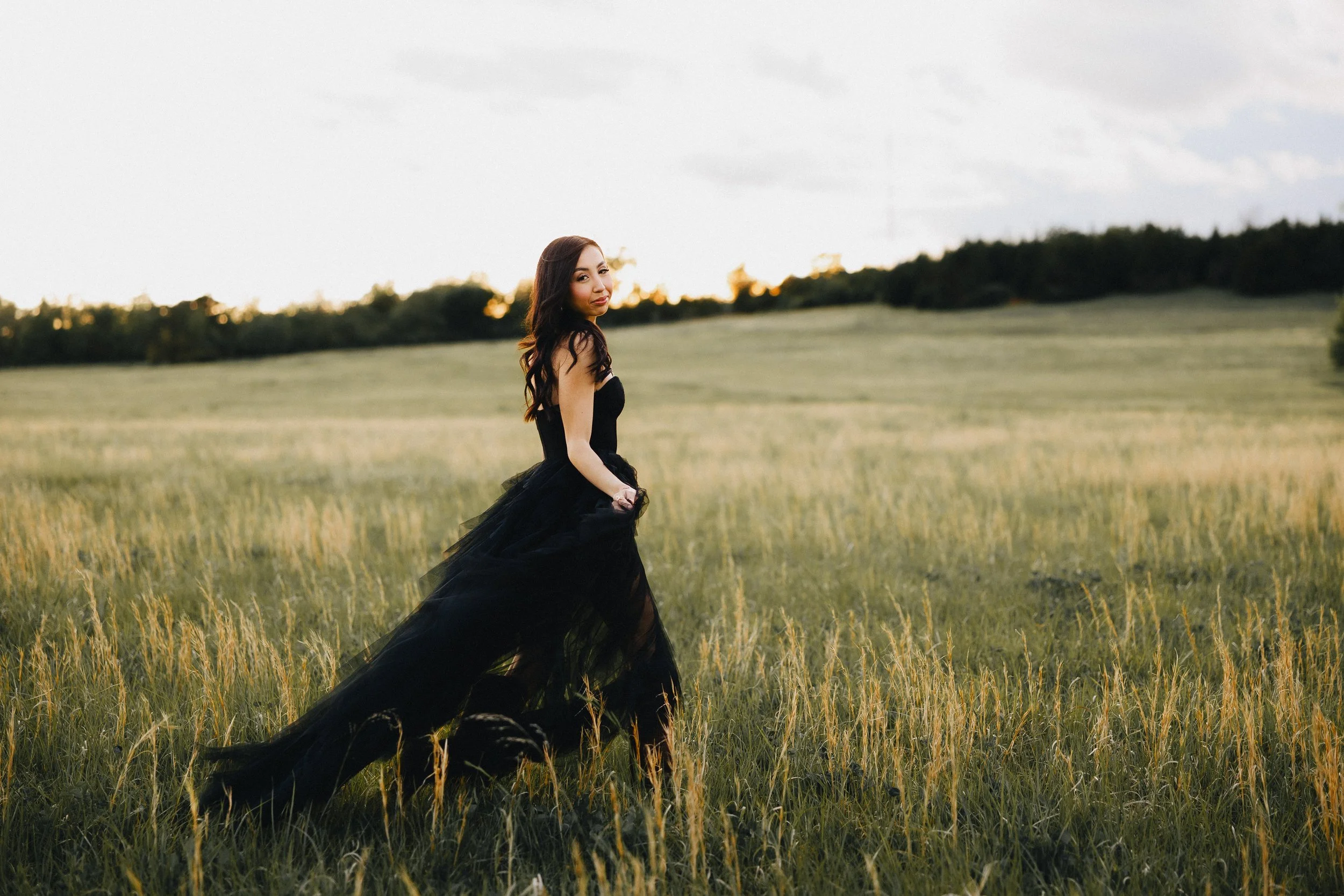 A woman in a long black dress standing in a grassy field during sunset.