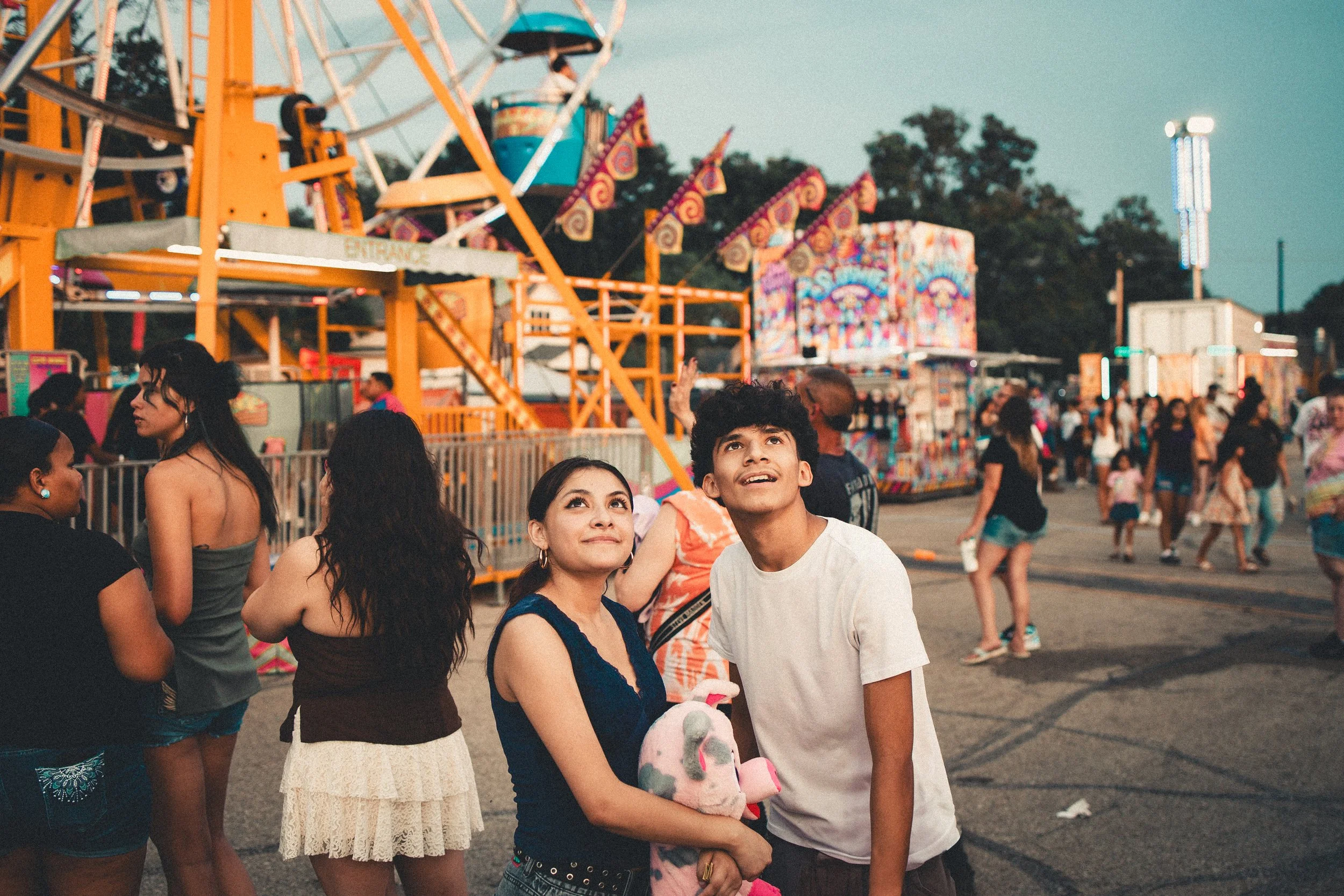 People at a carnival with a Ferris wheel and amusement rides in the background, some looking up and smiling. Shot at Fiesta Topeka.