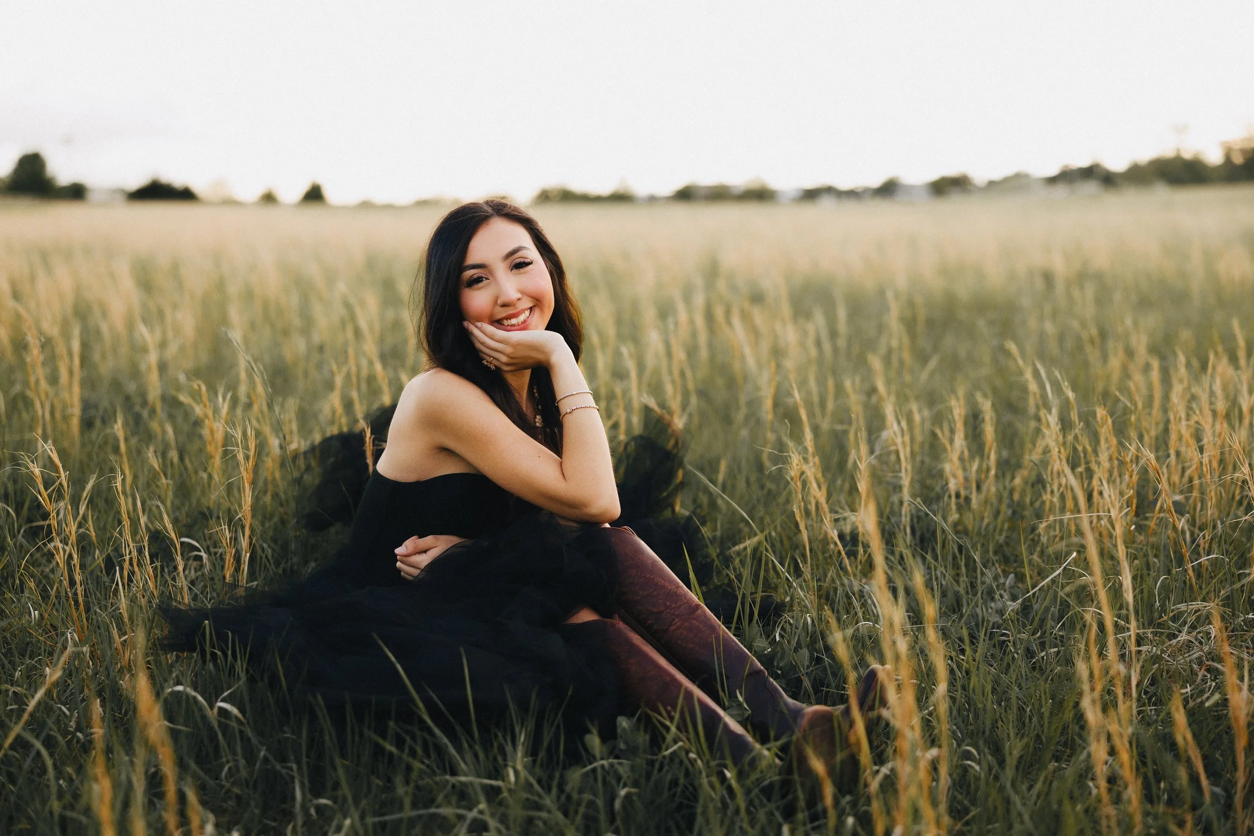 A young woman with dark hair, wearing a black dress, sitting on grass in a field, smiling and resting her chin on her hand.