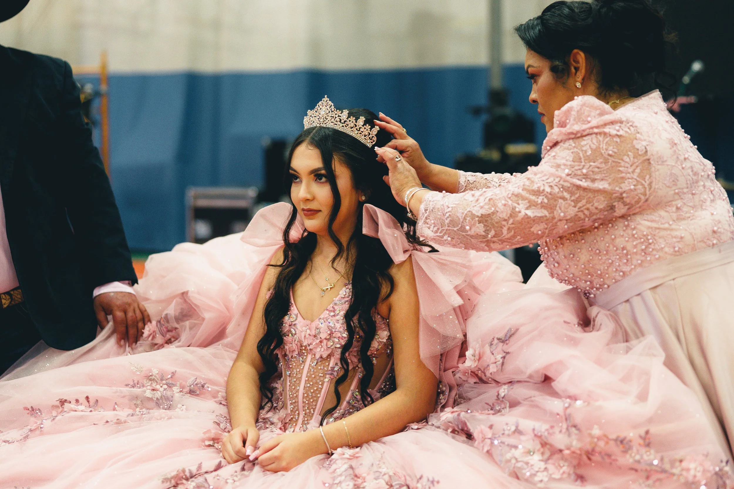 A young woman in an elaborate pink ball gown is having a tiara placed on her head by an older woman in a pink embroidered dress. They are indoors, possibly at a special event or celebration.