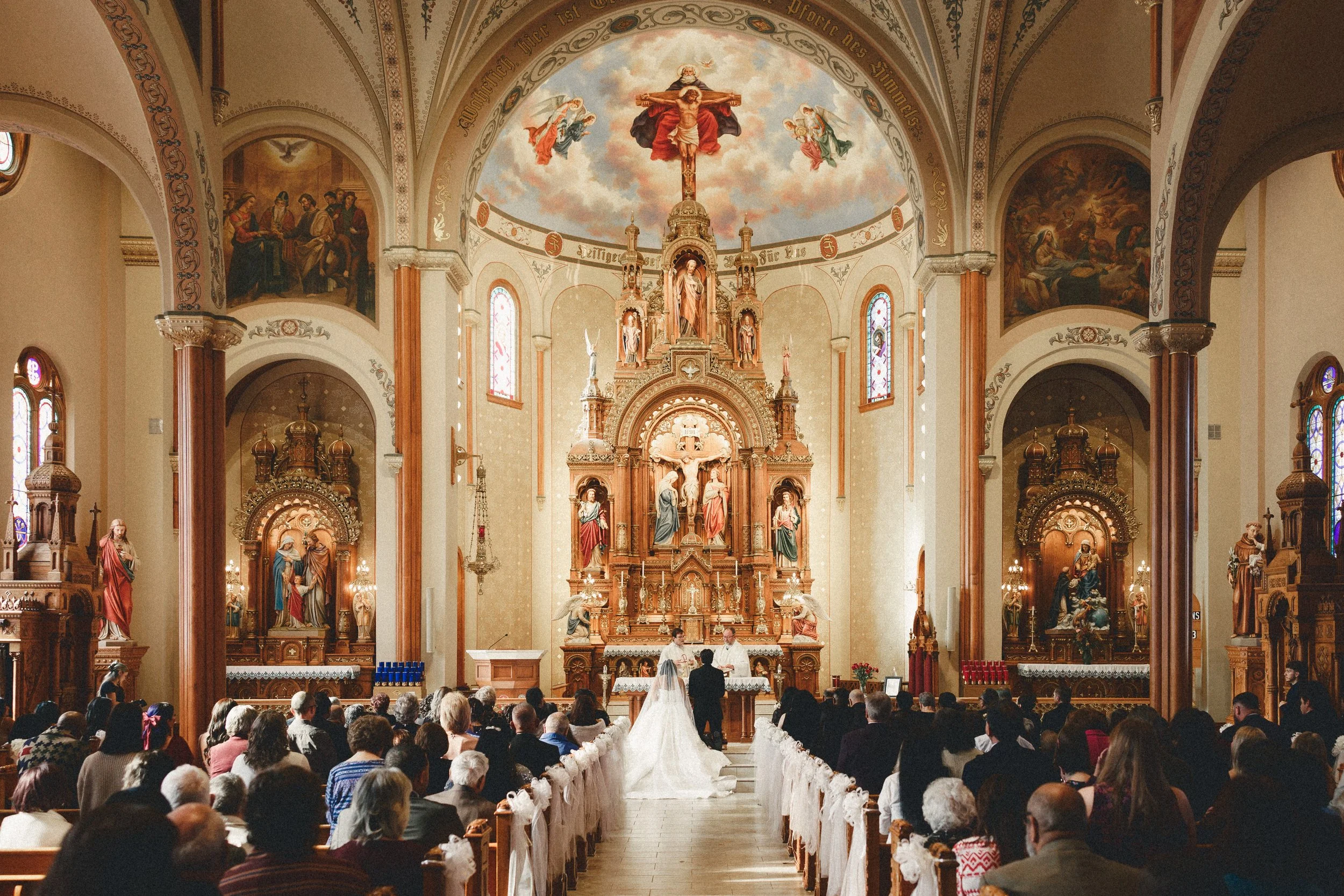 A wedding ceremony taking place inside a church with an ornate altar and religious paintings. The bride and groom stand at the altar facing a priest, with the congregation seated on either side.