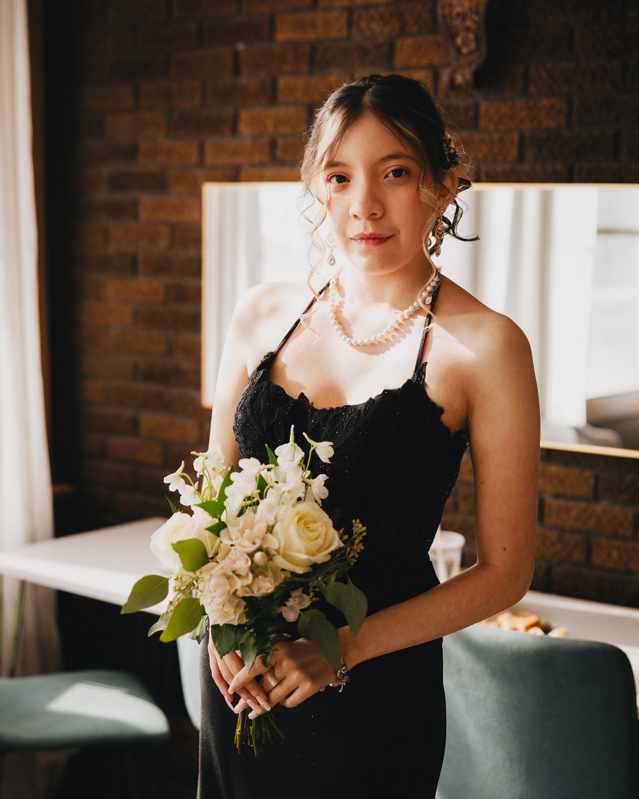 A young woman in a black dress holding a bouquet of white roses and greenery, standing indoors near a window with curtains and exposed brick wall. Shot at Topeka Kansas venue The Beacon.
