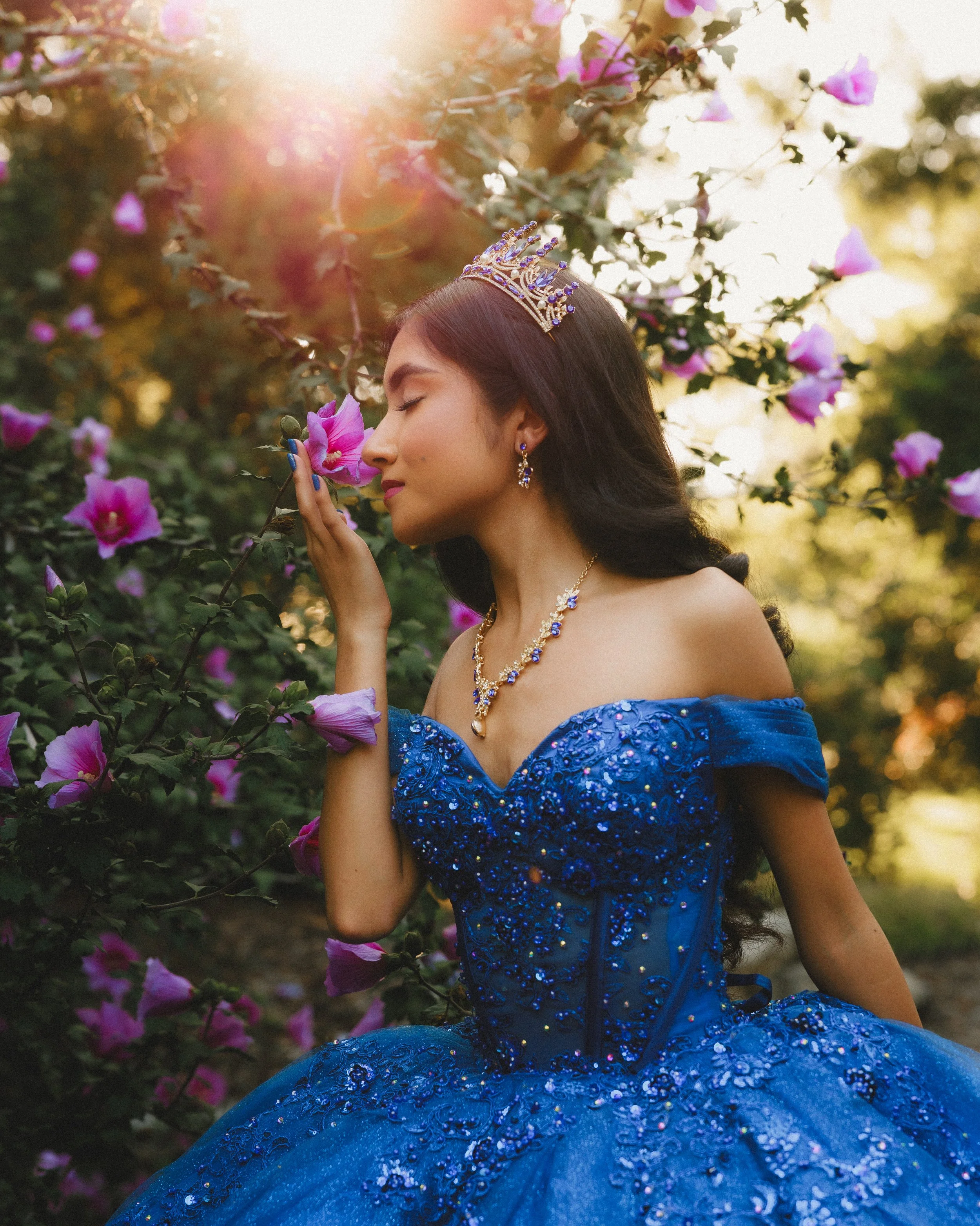 A woman in a blue, sequined, off-shoulder gown with matching jewelry and a crown, standing among pink flowers and holding a flower close to her nose with her eyes closed.
