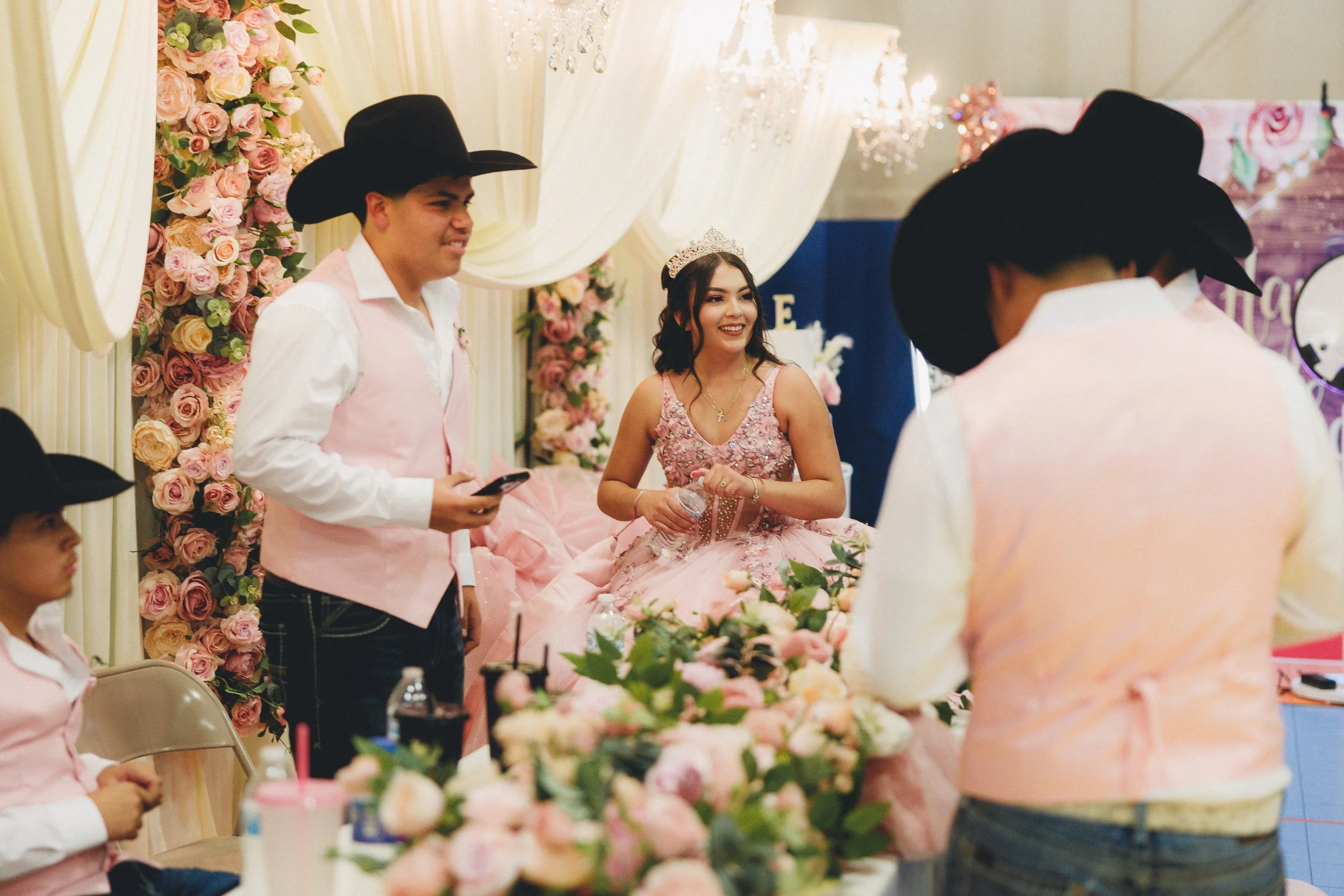 A young woman in a pink dress with a tiara and necklace, smiling and holding a water bottle, surrounded by young men in cowboy hats and pink vests at a decorated party with pink roses and floral arrangements.