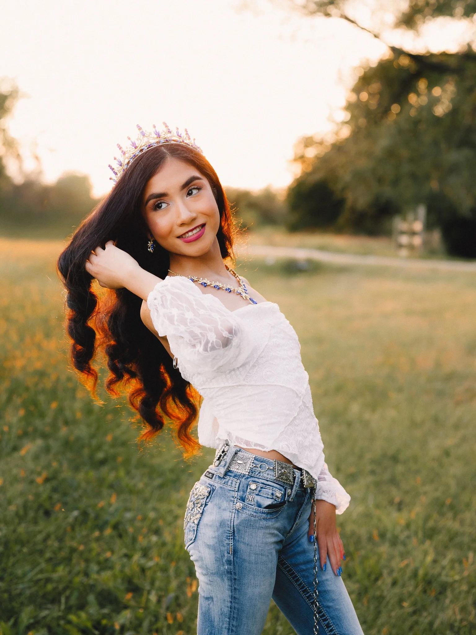 A young woman with long dark hair wearing a crown, a white lace top, and blue jeans poses outdoors at sunset, smiling and touching her hair.