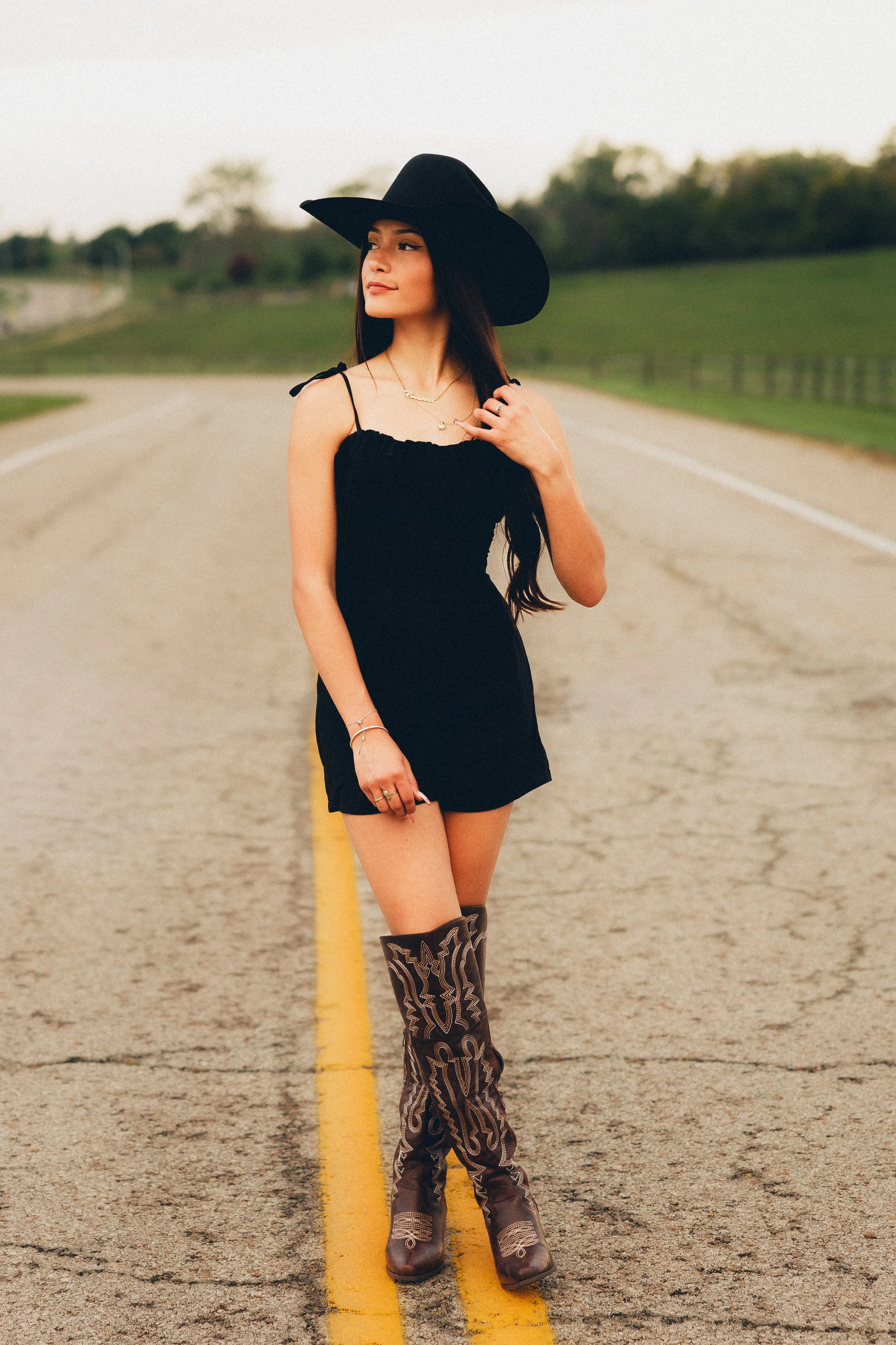 A young woman standing on a rural road wearing a black dress, large black hat, and cowboy boots.