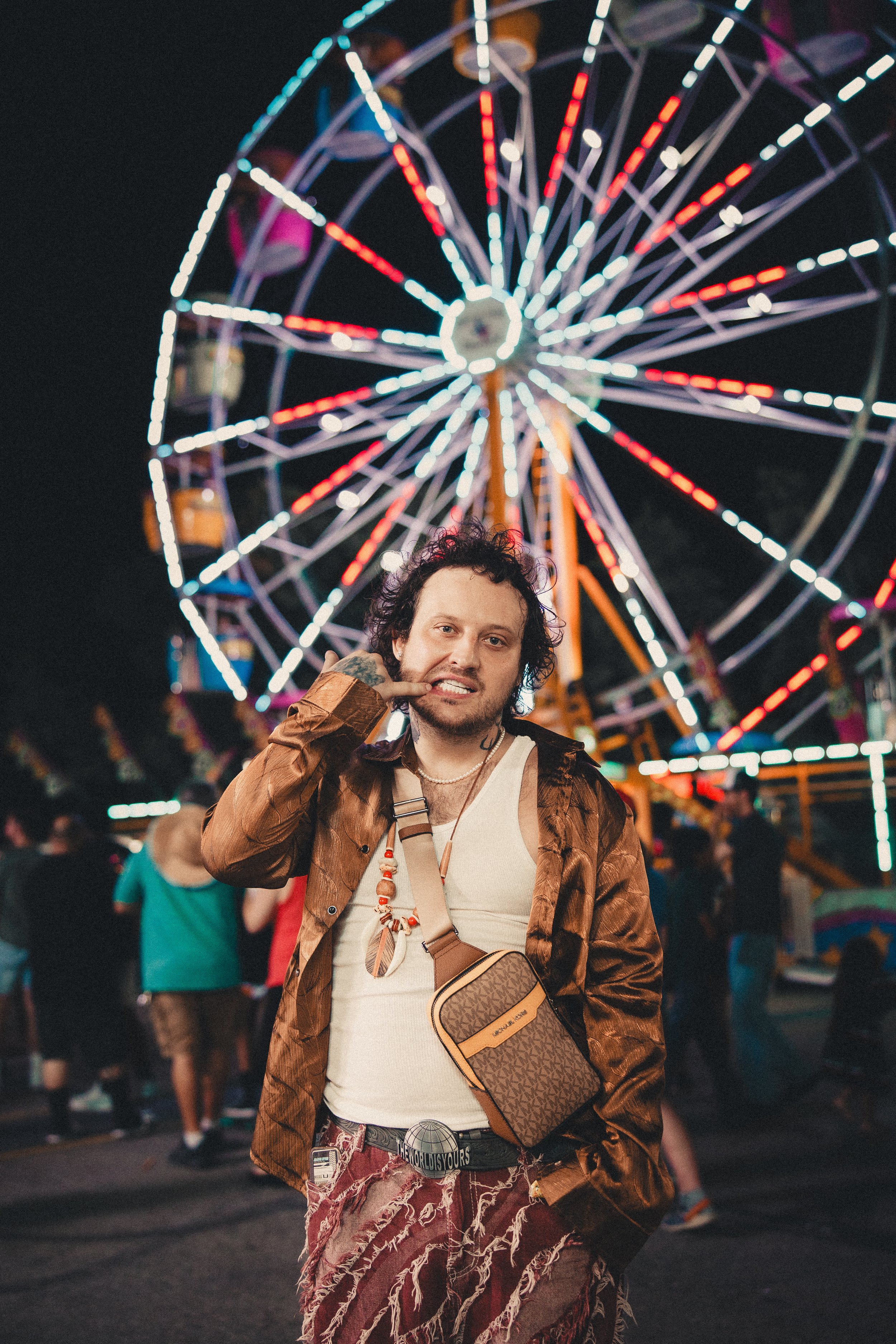 A person with curly hair in front of a Ferris wheel at night, making a gesture towards their face with their hand. The background shows bright, colorful lights from the amusement park. Shot at Fiesta Topeka.