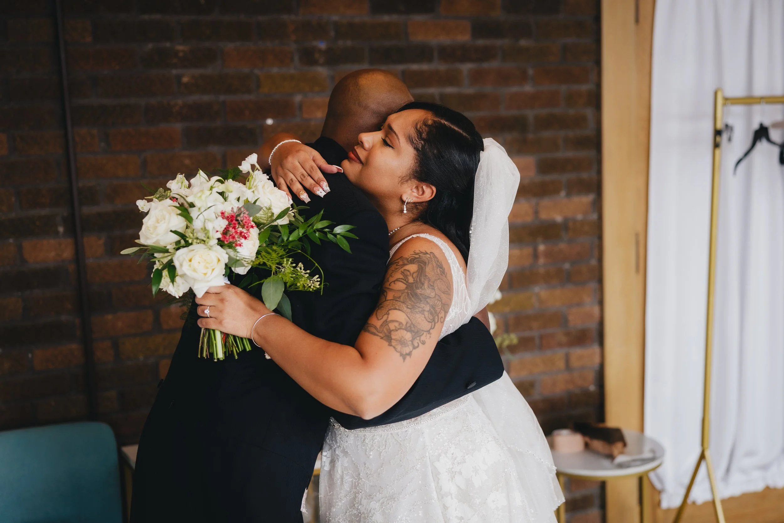 A bride and groom hug, with the bride holding a bouquet of white and pink flowers, against a brick wall. Shot at Topeka Kansas venue The Beacon.