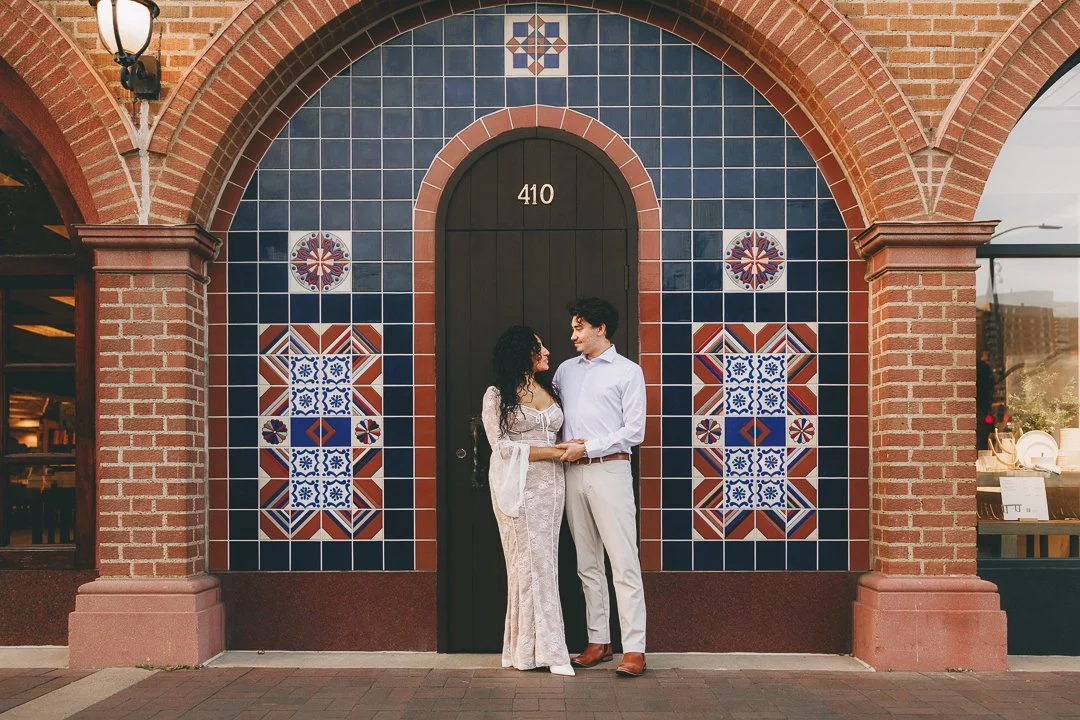 A couple standing hand in hand outside a building with a decorative tile wall background featuring red, blue, and white patterns, brick archways, and a black door with the number 410. Shot at Kansas City, Country Club Plaza.