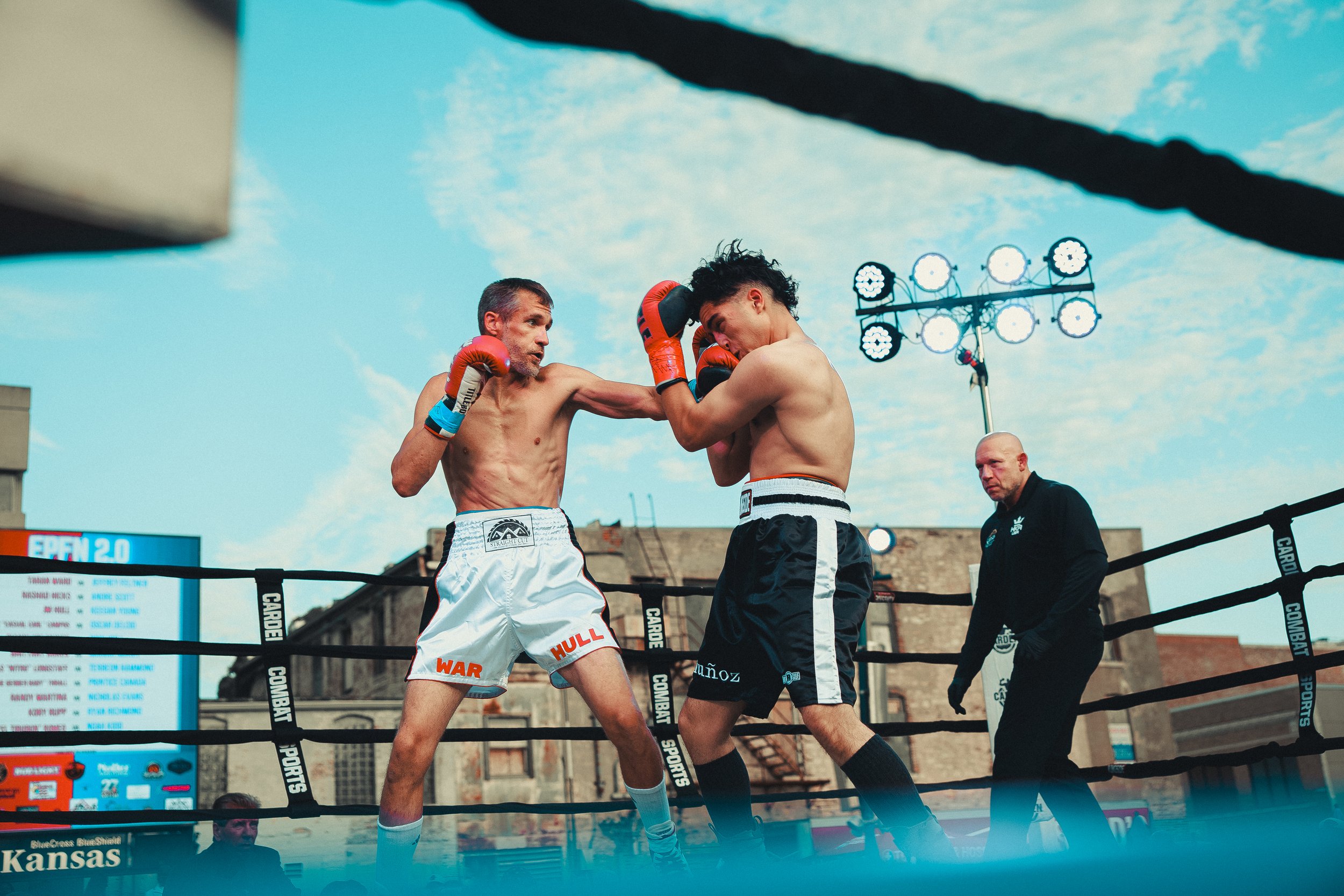 Two shirtless male boxers are sparring in a boxing ring outdoors, with one delivering a punch to the other's face, and a referee watching nearby. The scene is set in an urban environment with buildings and spotlights in the background. Shot at Evergy