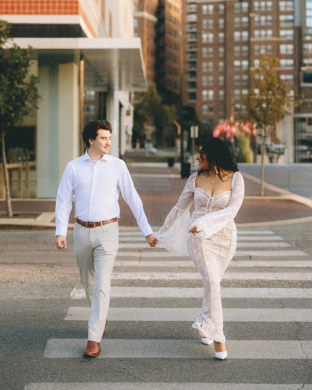 A young man and woman holding hands, walking across a city crosswalk in the late afternoon or early evening. The man is dressed in a white shirt and light-colored pants, while the woman wears a white dress with long sleeves and white heels. They are 