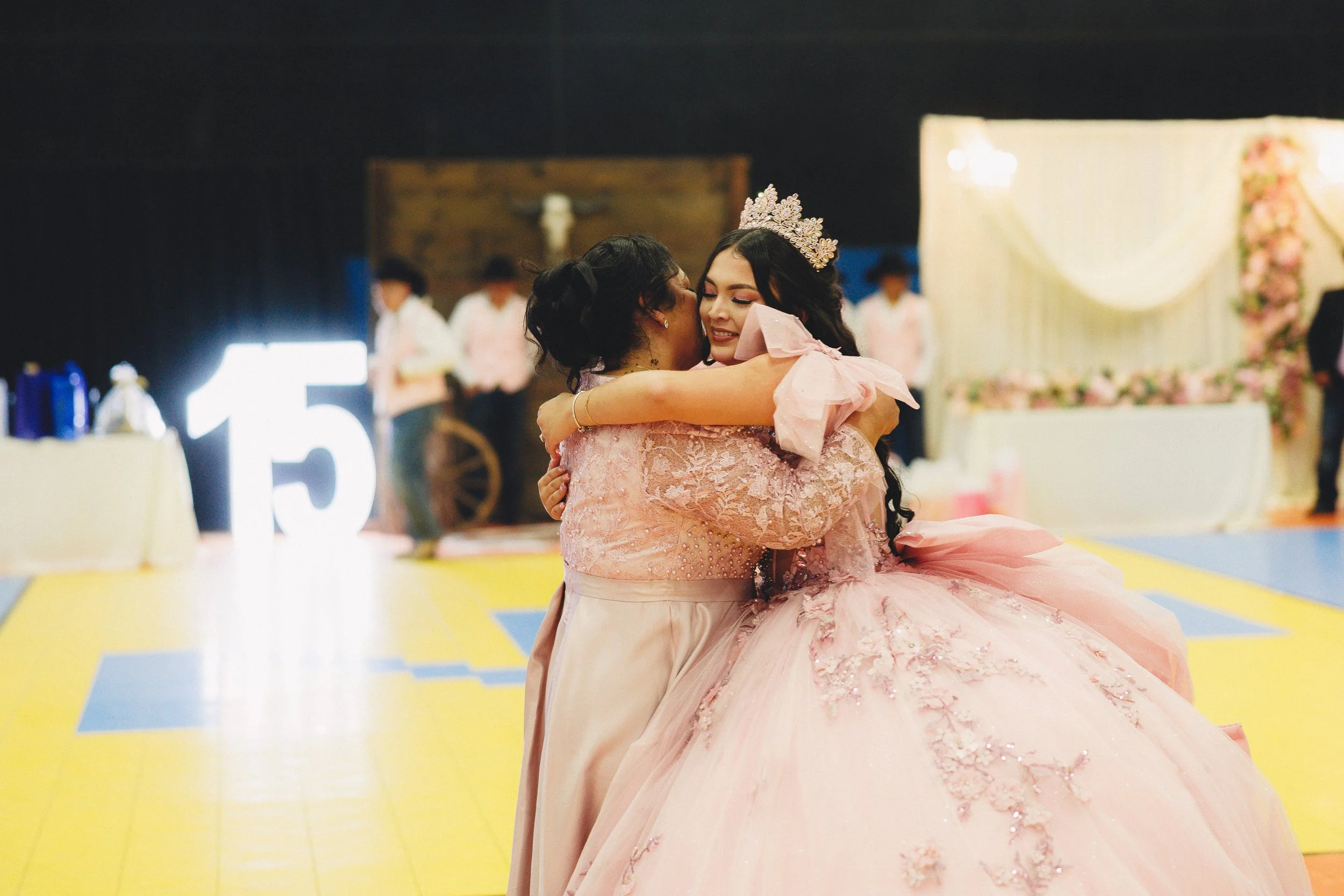Two women hugging at a quinceañera celebration, with one wearing a pink ball gown and tiara, and the other in a pink dress, on a decorated dance floor in the background.