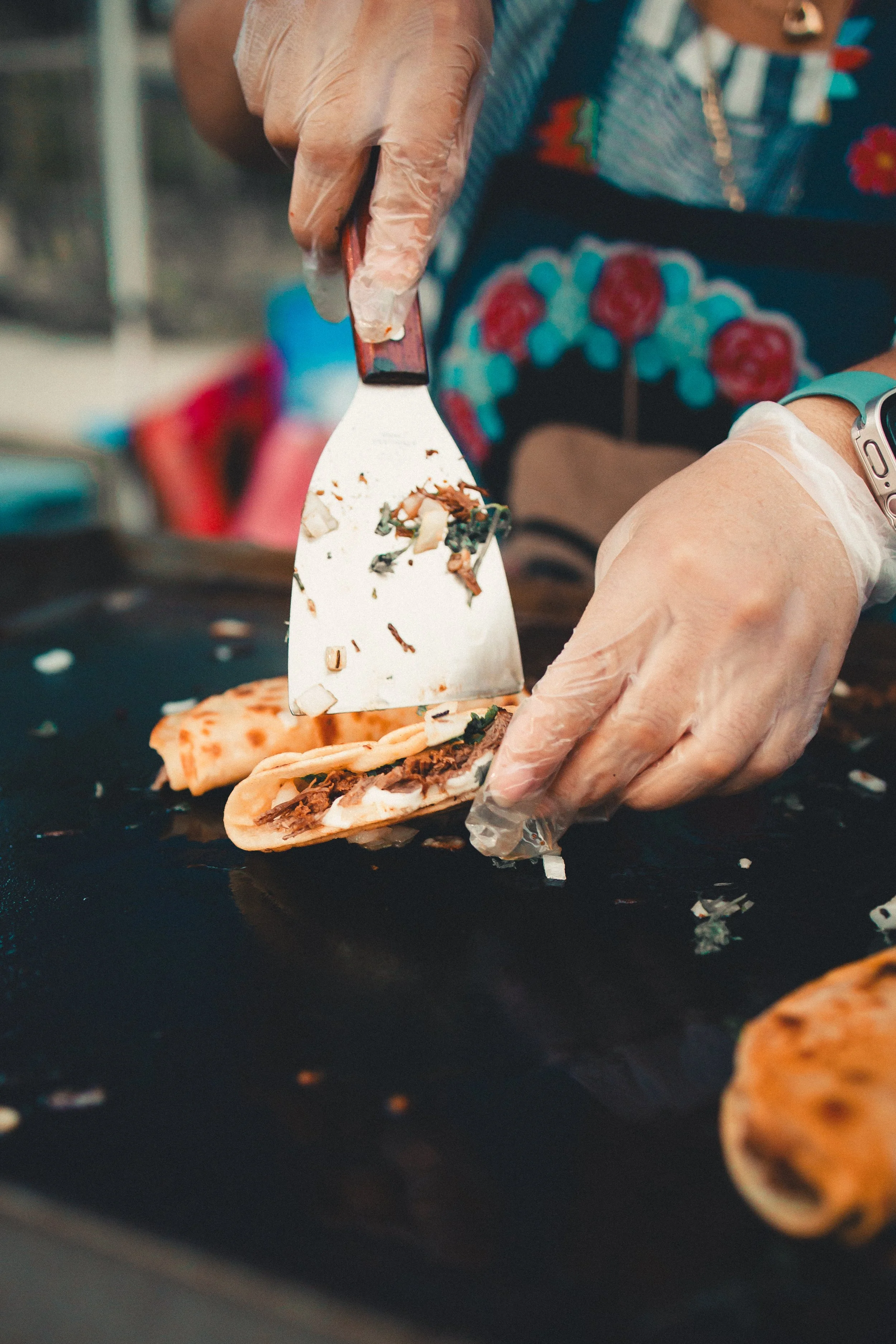 Someone wearing gloves preparing a taco on a griddle, with colorful clothing and apron. Shot at Fiesta Topeka.