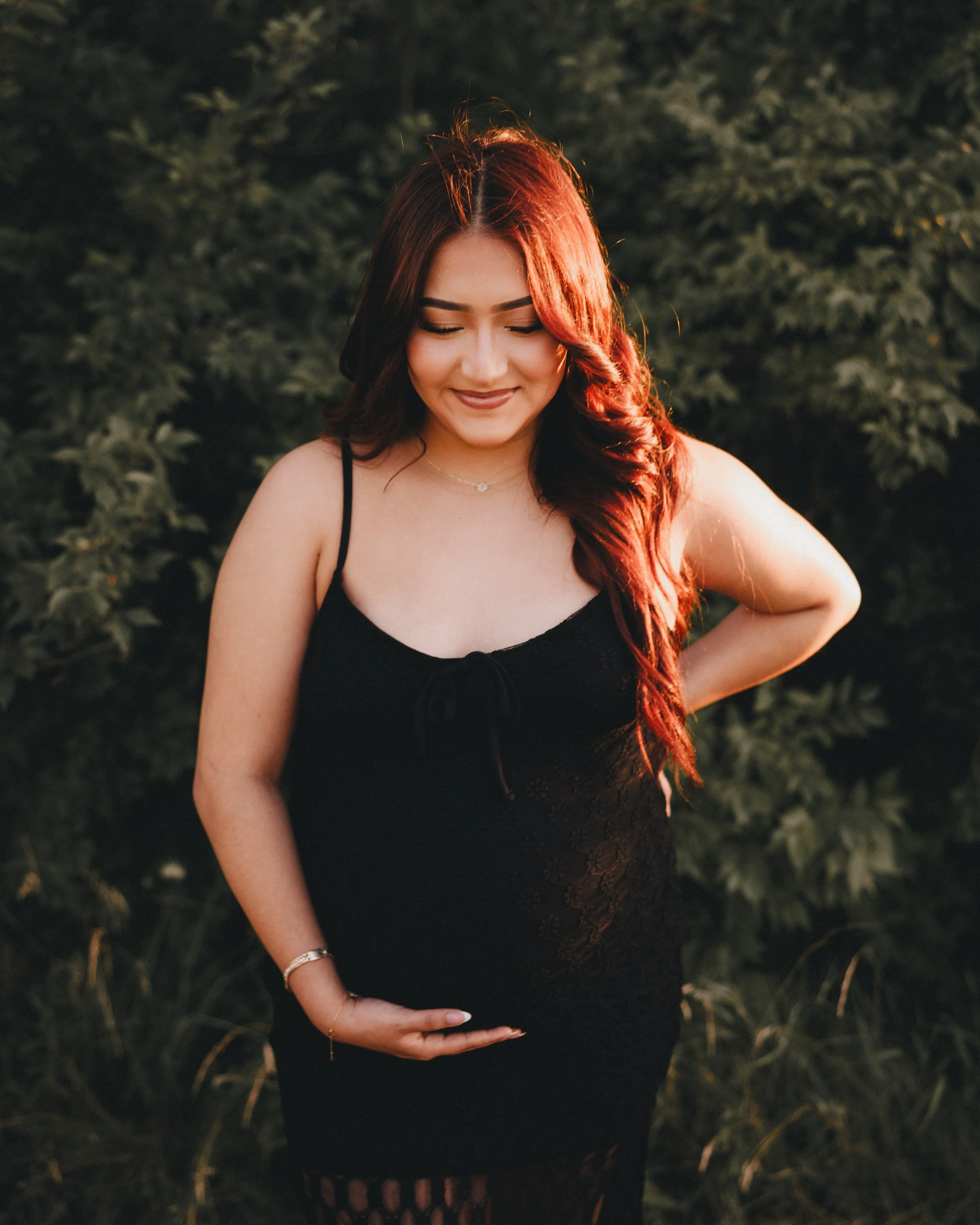 A pregnant woman with long red hair wearing a black dress standing outdoors in front of green foliage, smiling with eyes closed and one hand on her belly.  Shot in Topeka, Kansas. 