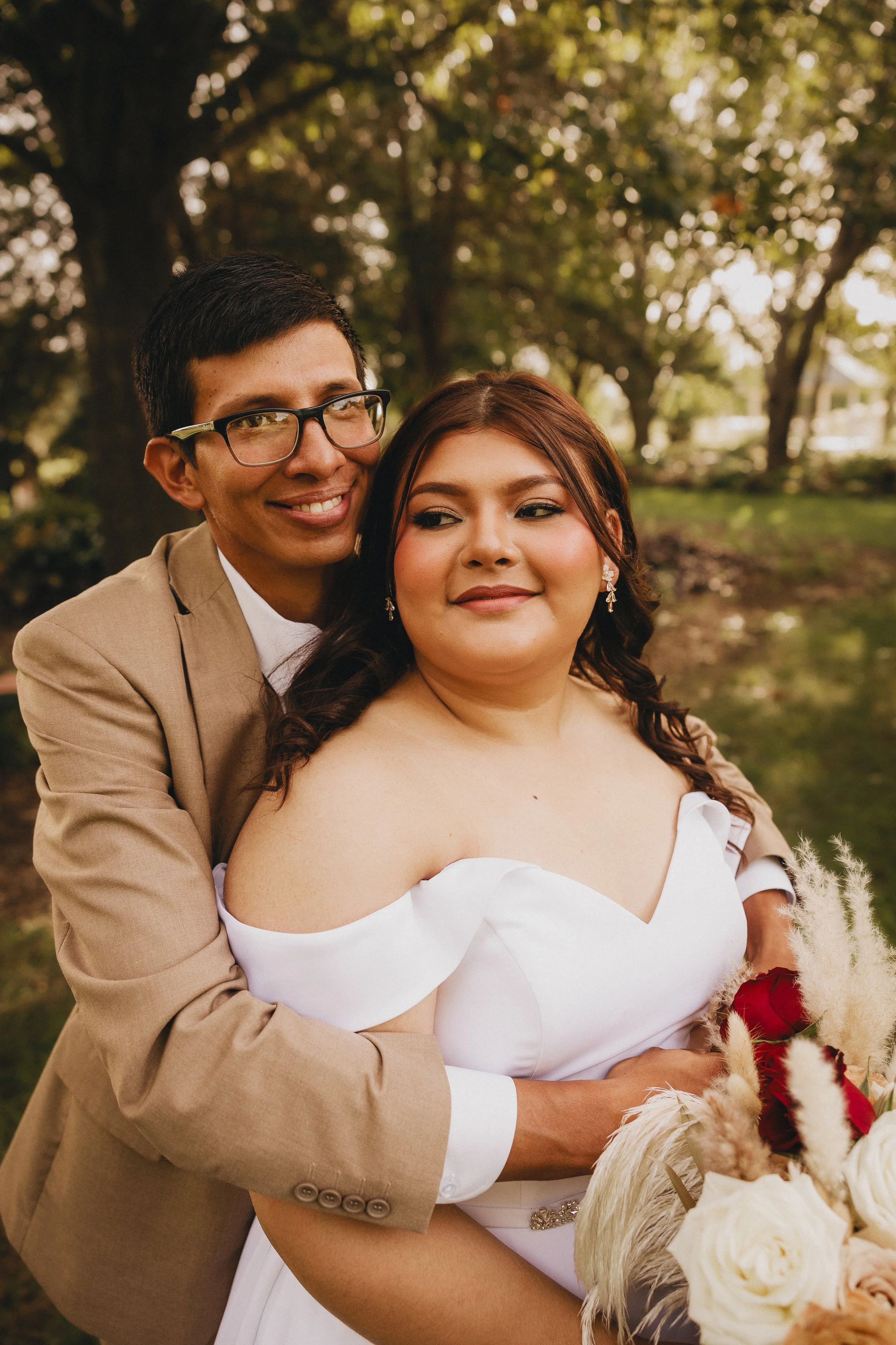 A couple at a wedding or special event outdoors, with the man in a beige blazer and glasses, and the woman in a white off-shoulder dress holding a floral bouquet, surrounded by trees.