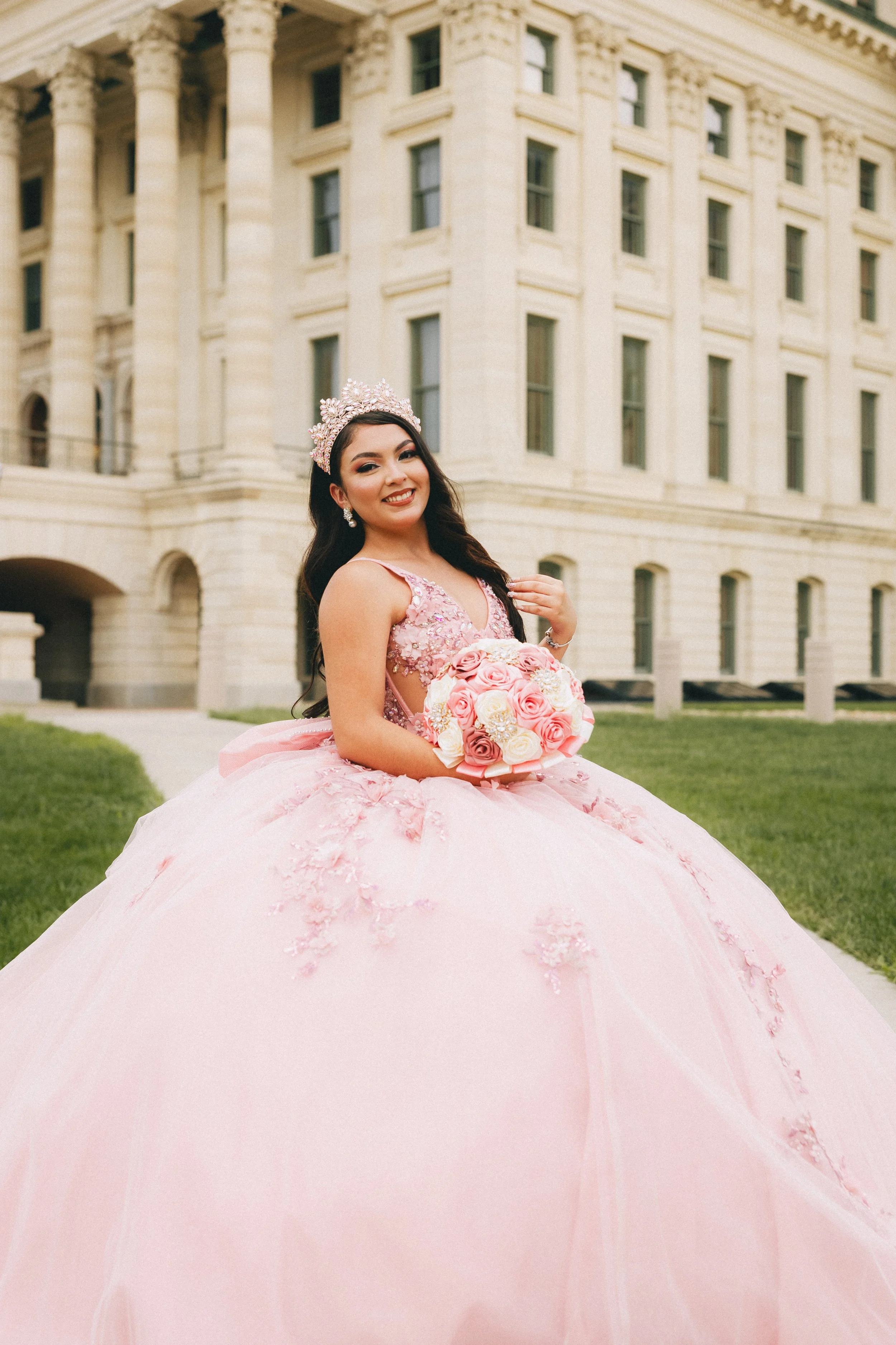 Young woman in a pink quinceañera dress, wearing a tiara, holding a bouquet of pink and white roses, standing in front of a historic building. Shot in Topeka, Kansas.