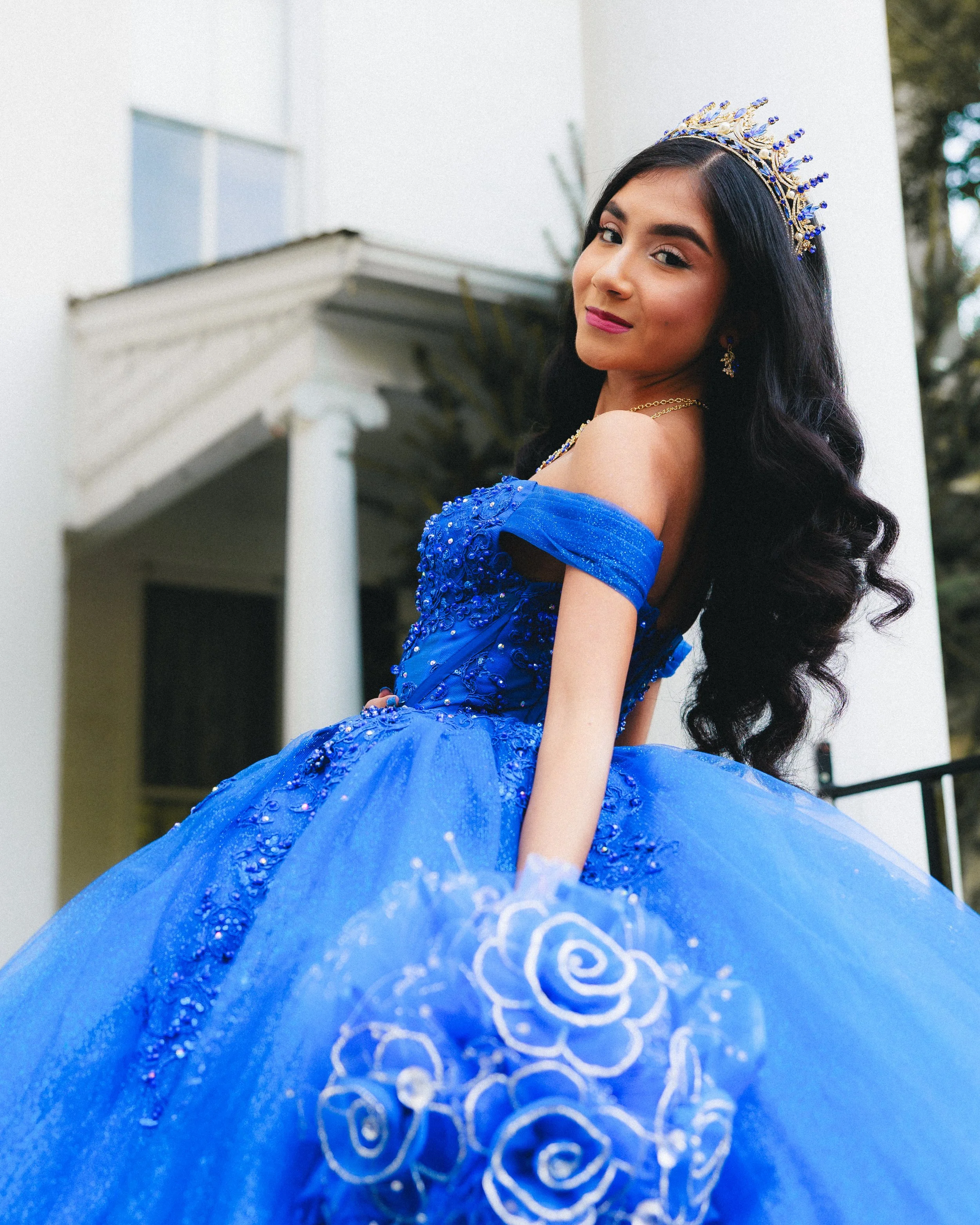 Young woman in a royal blue dress with floral embroidery, a tiara, and jewelry, posing outdoors in front of a white house with columns.