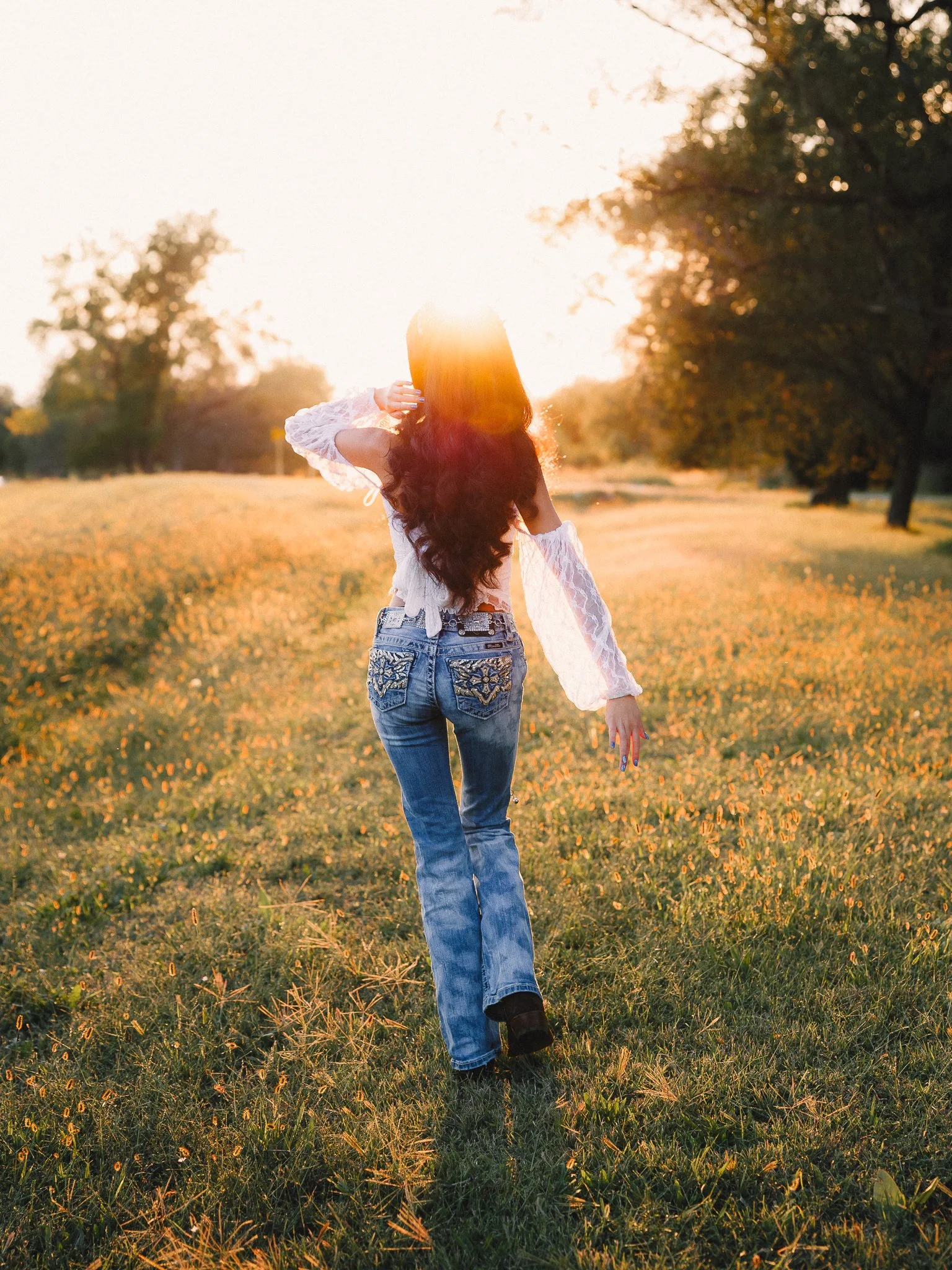Woman with long dark hair walking through a grassy field at sunset, wearing a white lace top and blue jeans.