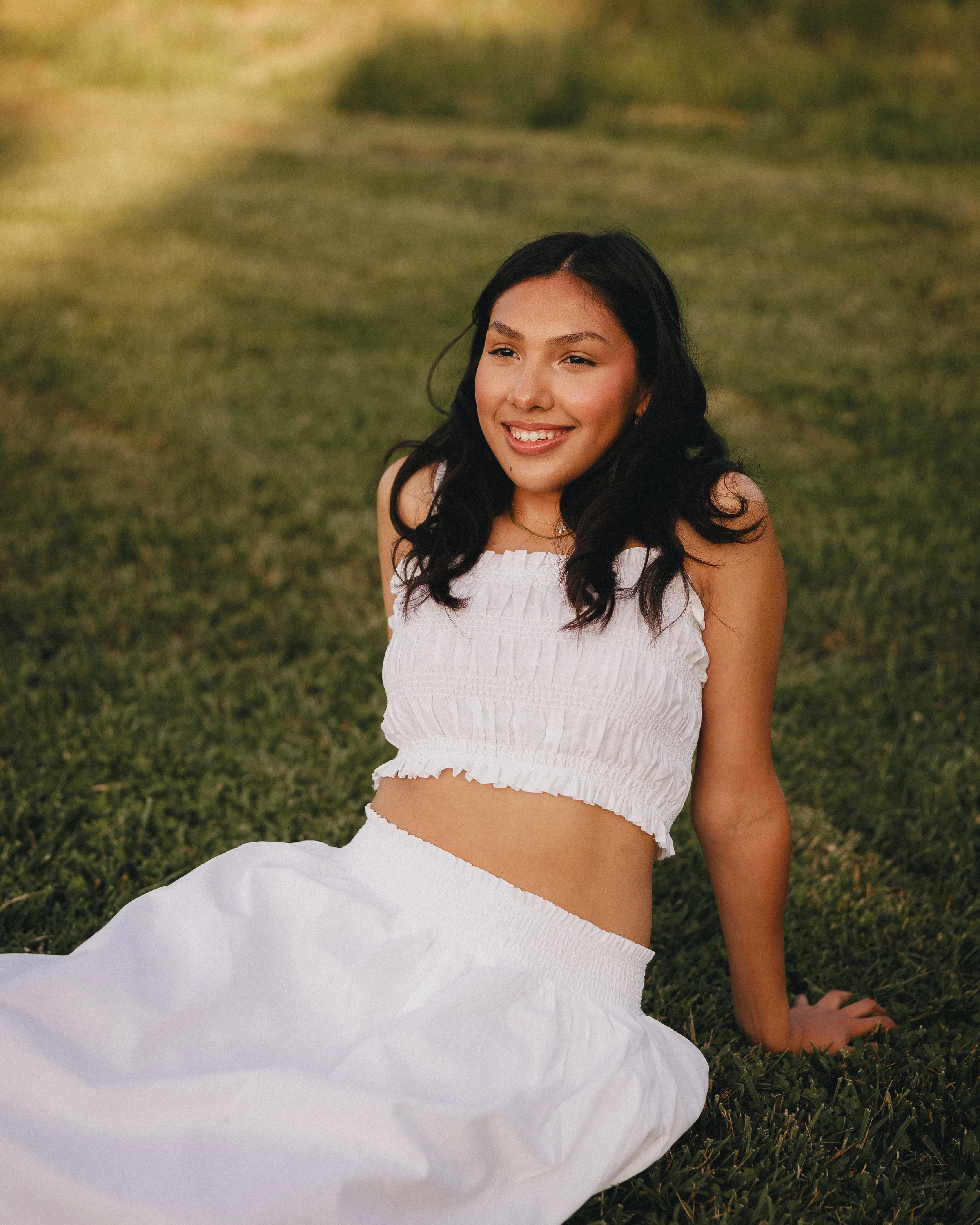 Young woman sitting on grass outdoors, smiling, wearing a white sleeveless top and white skirt, with dark wavy hair and natural makeup during the daytime.