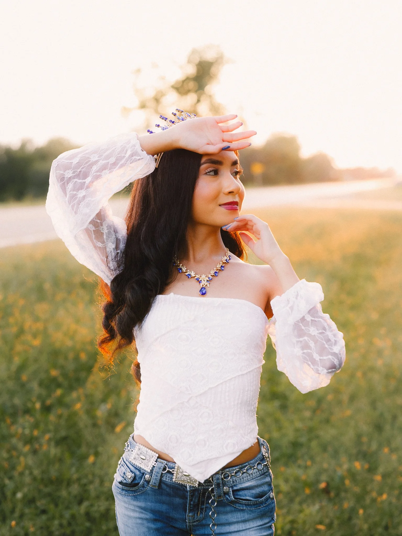 A woman with long dark hair posing outdoors at sunset, wearing a white off-the-shoulder top, jeans, a blue jewelry set, and a crown, with her hand shielding her eyes from the sun.