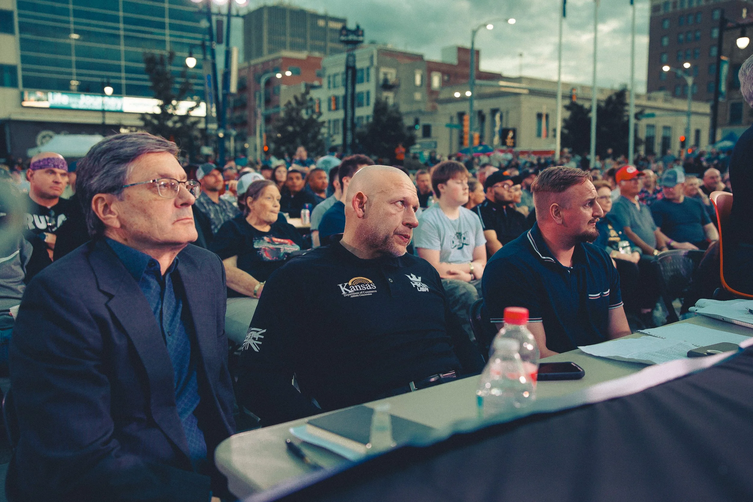 Audience at outdoor event, people seated at tables, city buildings in background, some looking attentive, with water bottles and papers on tables. Shot at Evergy Plaza in Topeka, Kansas. 