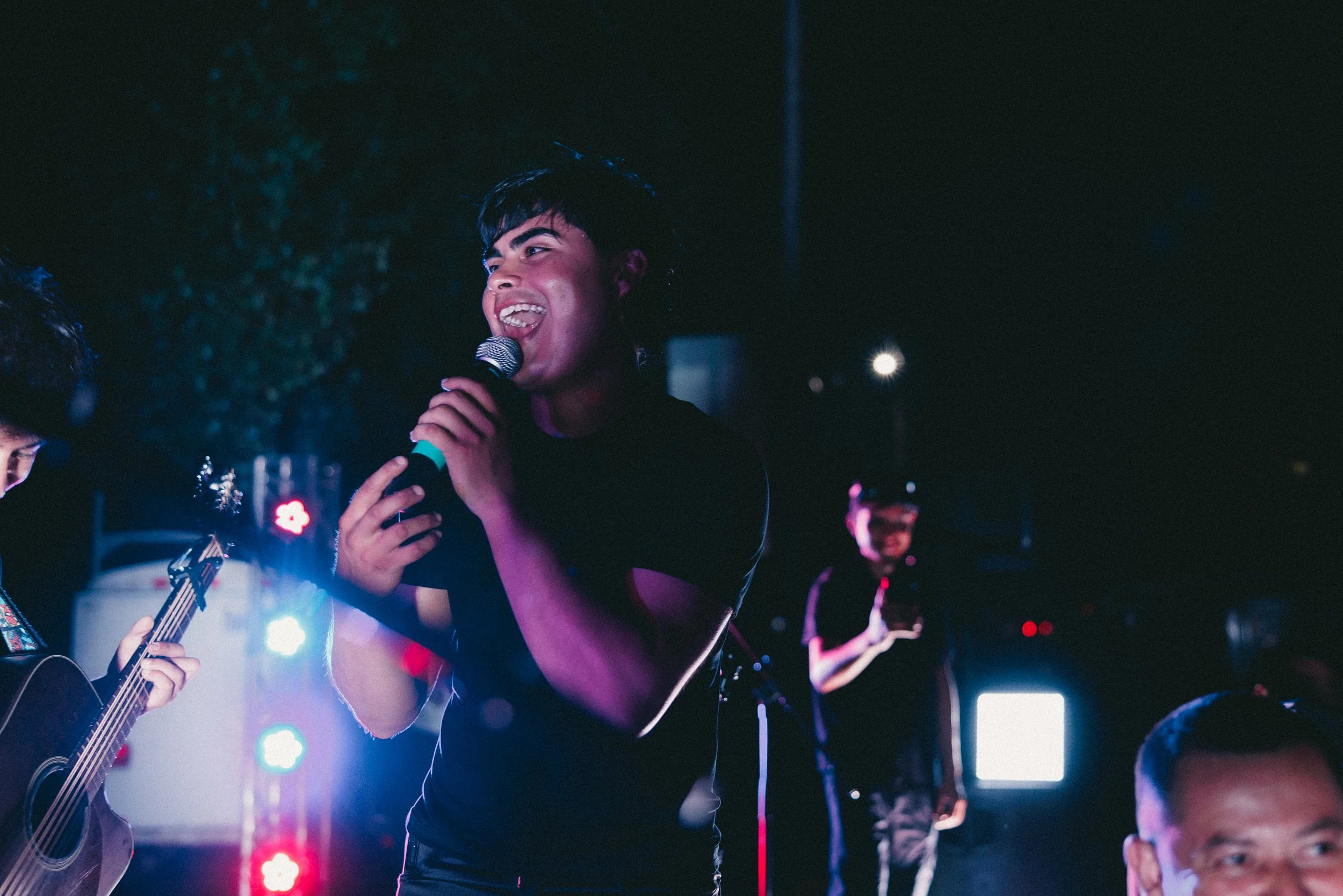 A young man with dark hair singing into a microphone on stage during a concert, with a guitarist on his left and another musician in the background, under colorful stage lights. Shot at Fiesta Topeka.