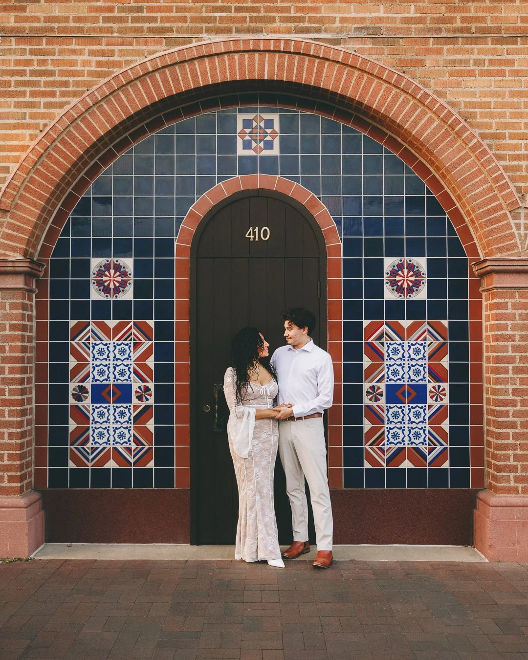A couple holding hands in front of a decorative brick and tile wall with the number 410 on a black door. Shot at Kansas City, Country Club Plaza.