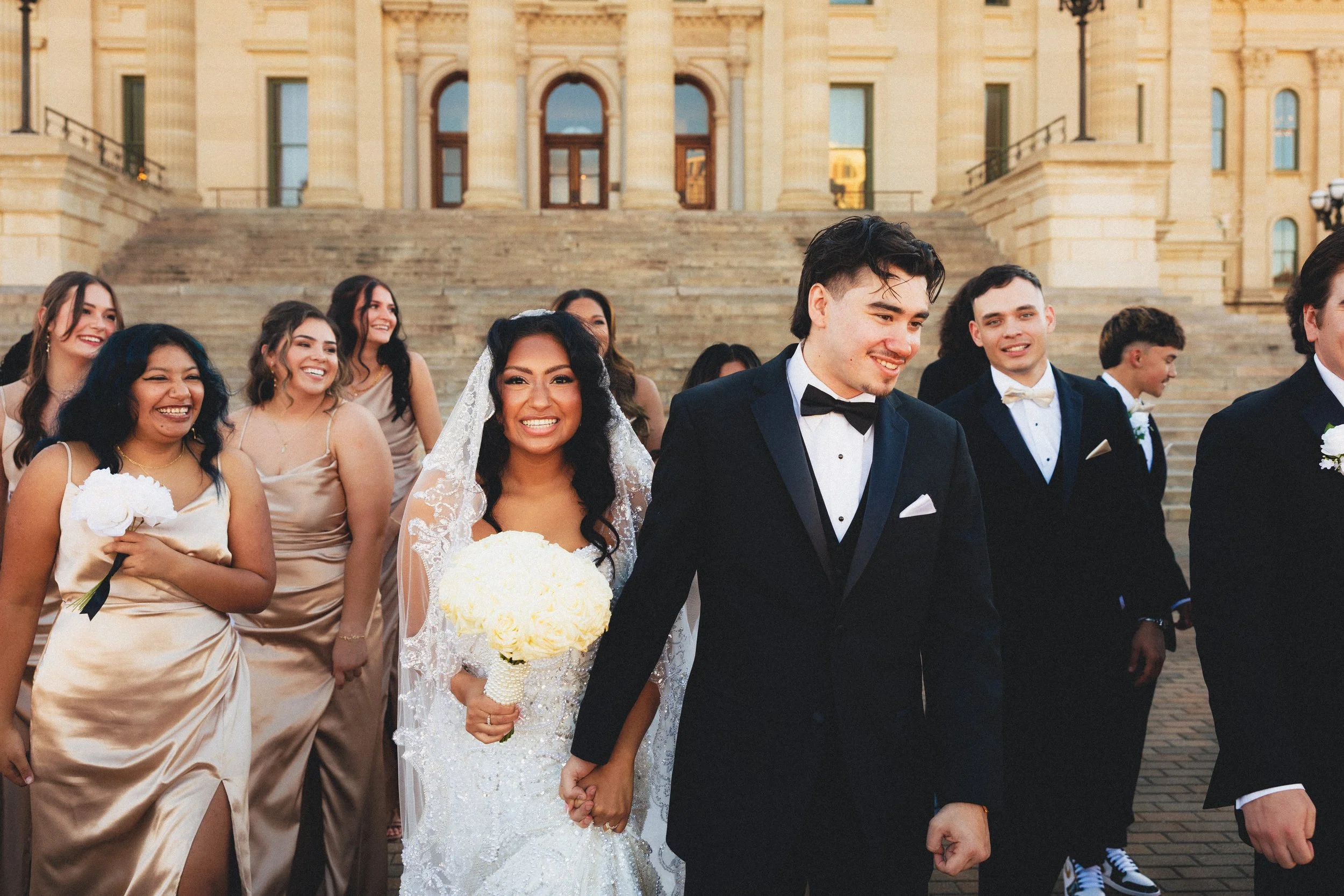 A wedding party standing outside a historic building with stairs, including a bride in a white gown and veil holding a bouquet, a groom in a black tuxedo, and several bridesmaids and groomsmen in formal attire, smiling and celebrating.